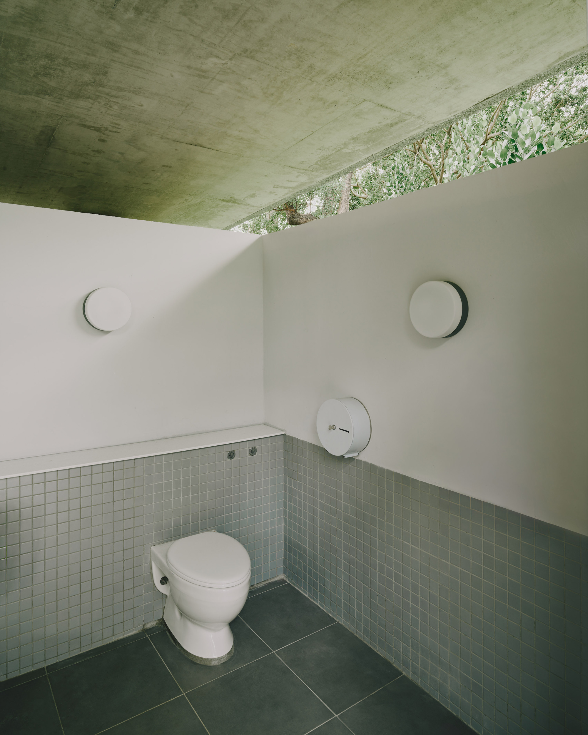 Public restroom interior with green mosaic tile wainscot and concrete ceiling opening to foliage above