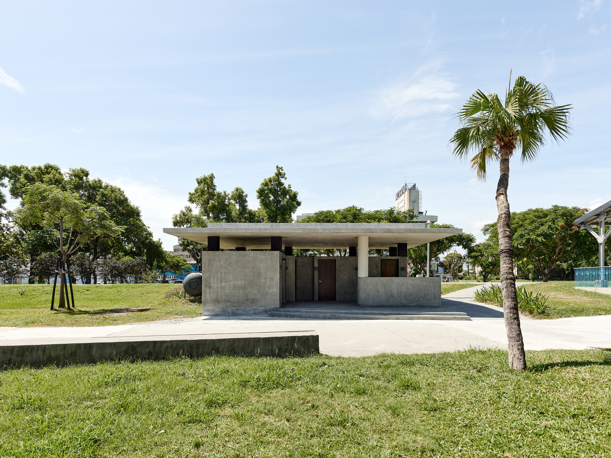 Front view of the concrete pavilion with flat roof surrounded by lawn and palm trees