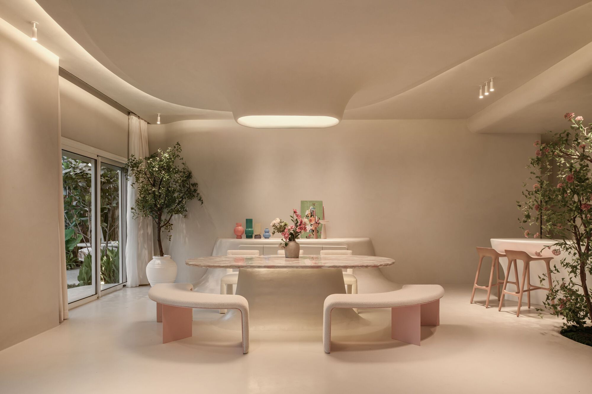 Dining area with a pale stone table, curved benches, and potted trees under soft ceiling light