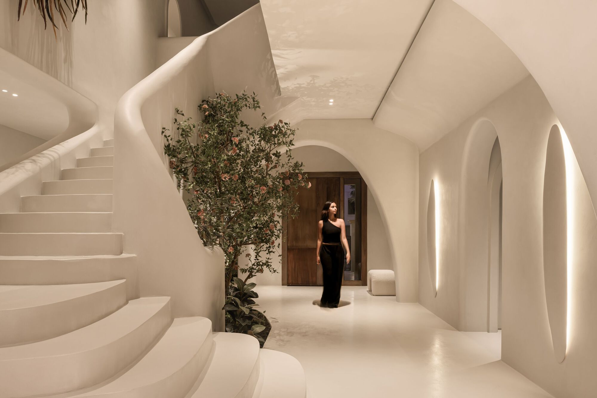 White curved entry hall with spiral staircase and arched doorways, a flowering tree and a visitor in evening light