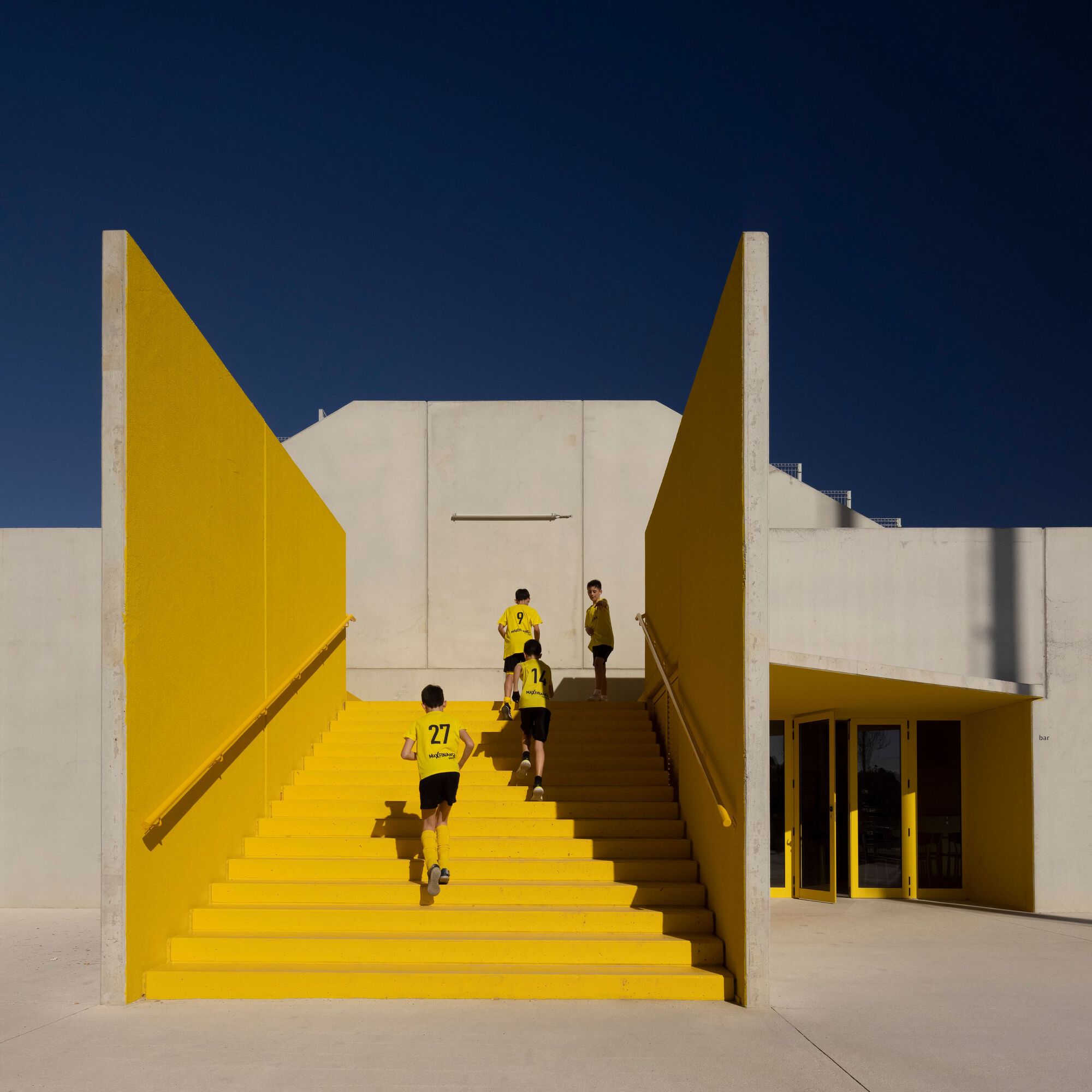 Yellow-painted exterior staircase flanked by angled walls leading to the upper level in bright sunlight