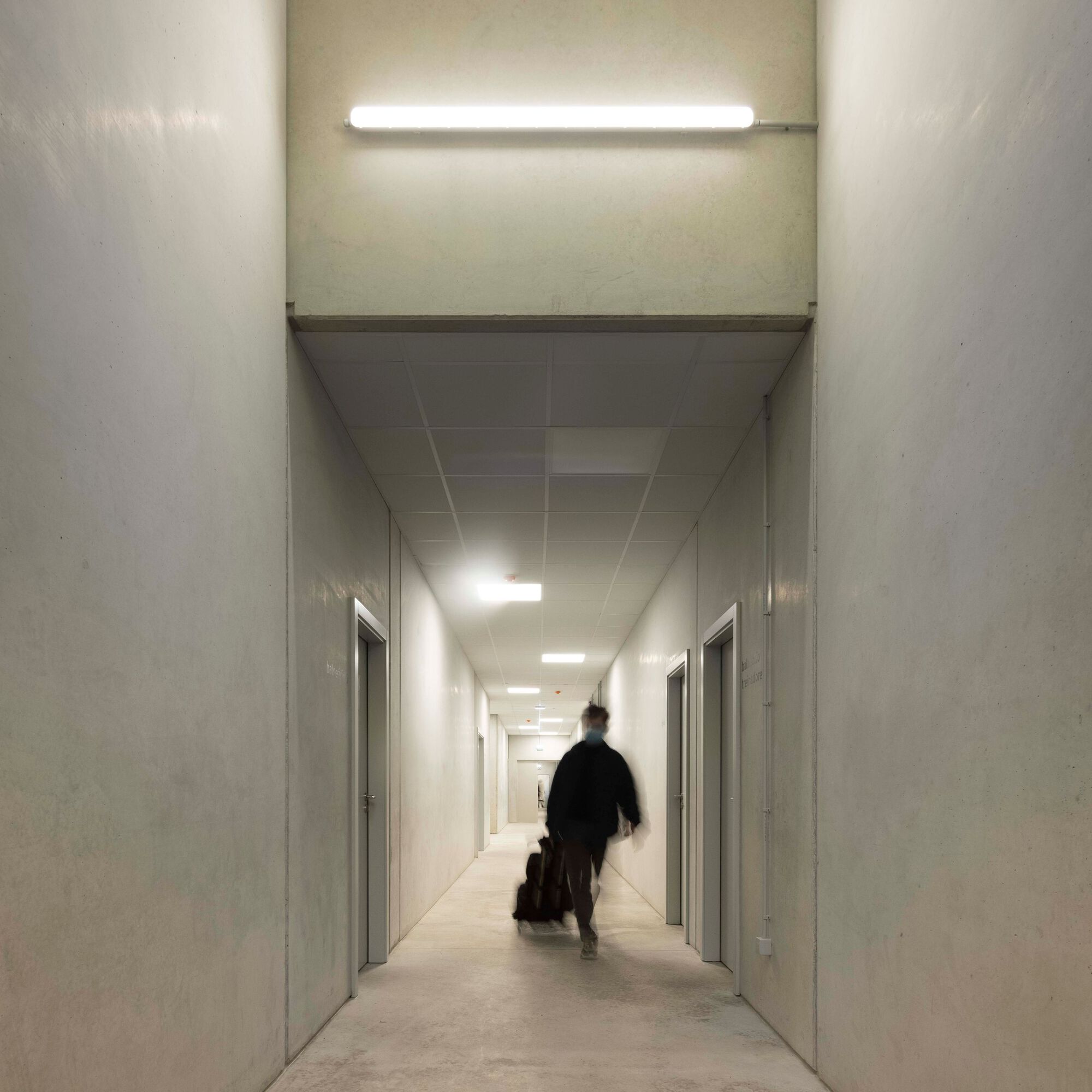 White corridor with fluorescent lighting and figure with luggage walking toward tiled ceiling terminus
