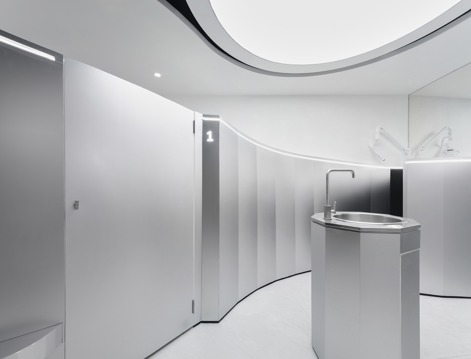 Cylindrical handwashing station with curved white cabinetry and toilet stall partitions under recessed lighting