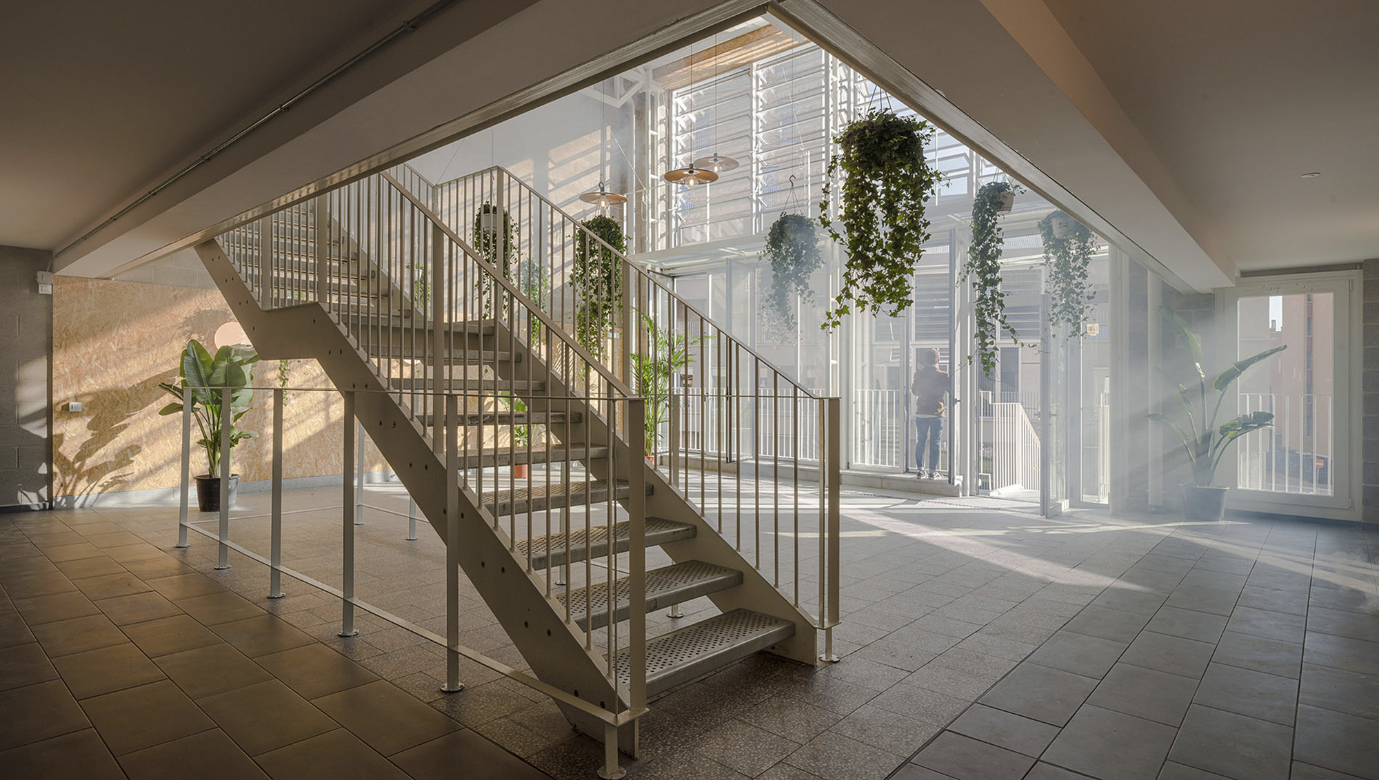Metal staircase with vertical rod railings rising through a skylit lobby with potted plants and sunlight