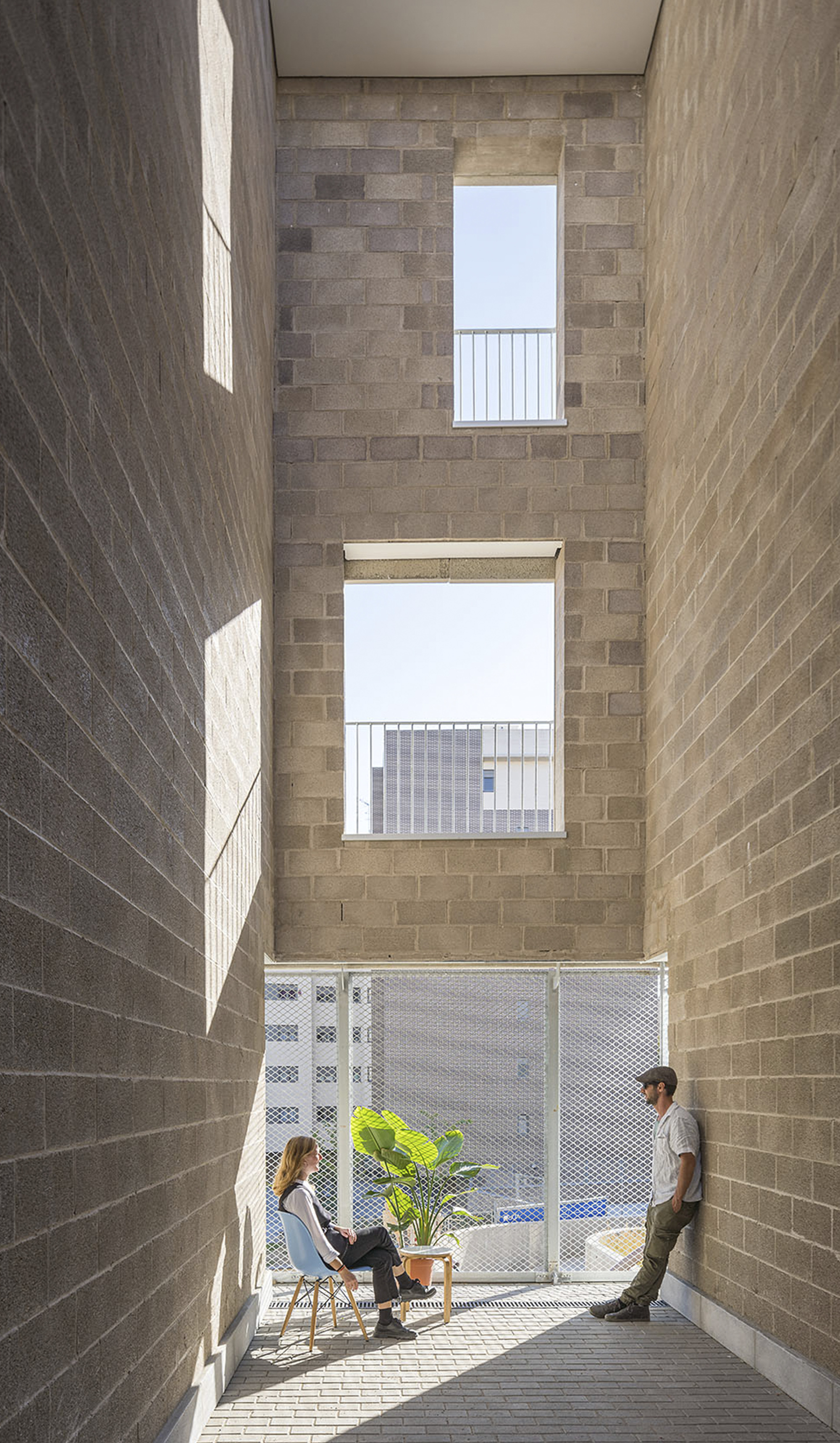 Narrow courtyard with brick walls, stacked window openings, and two people seated near a potted plant