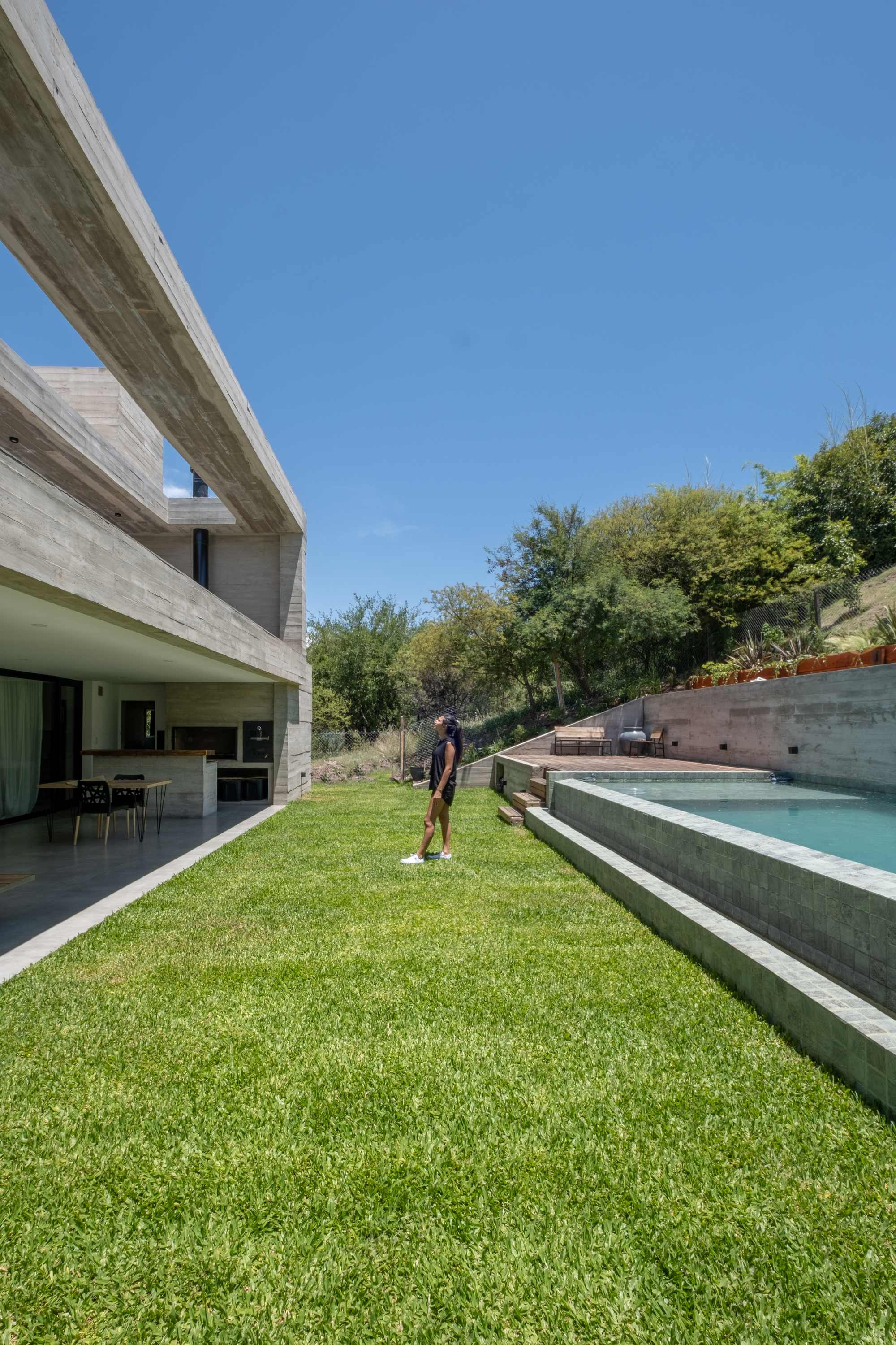Rear lawn sloping toward raised pool terrace with person walking under board-formed concrete canopy