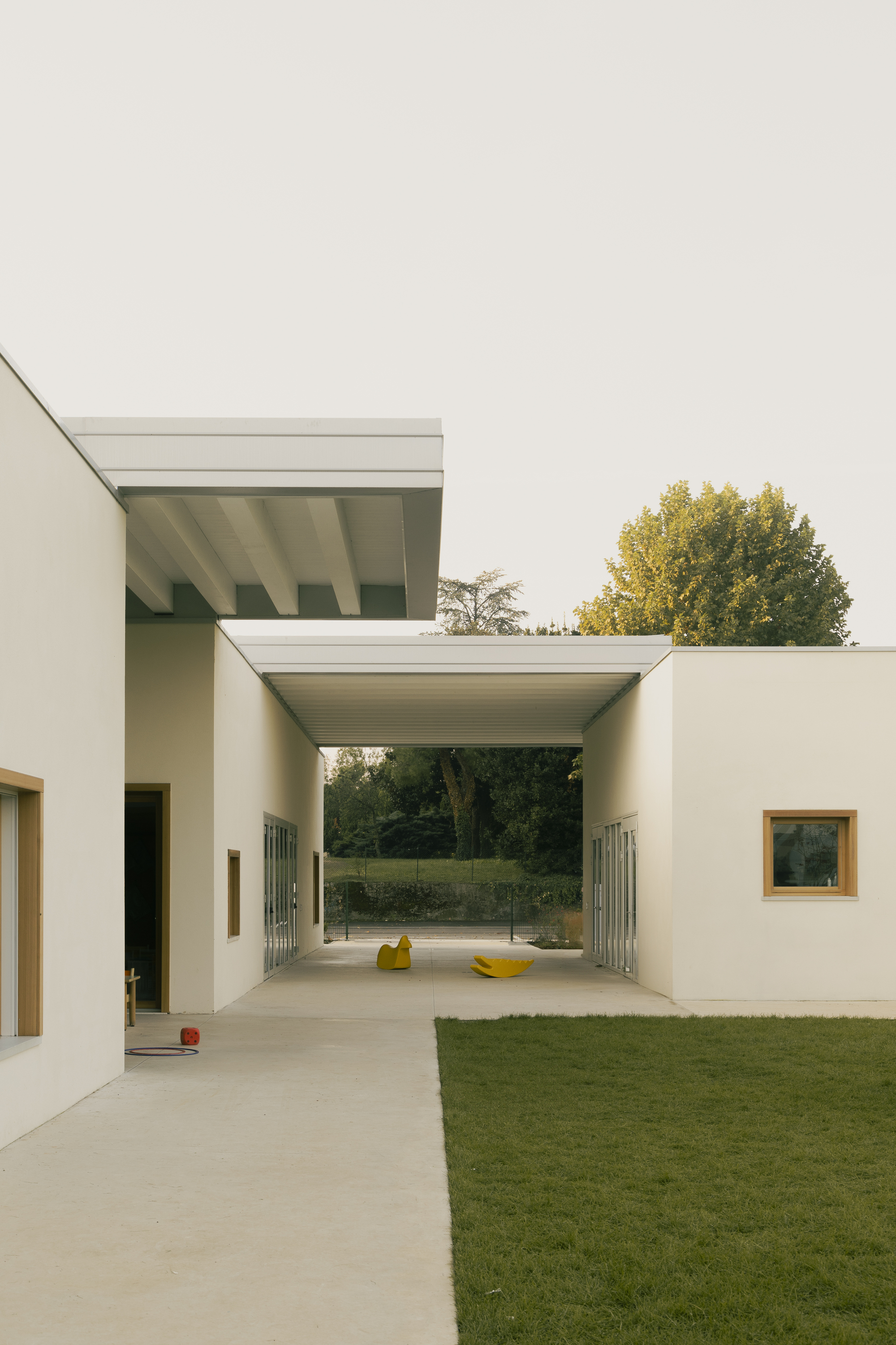 Covered passageway with exposed beam ceiling connecting white volumes across a courtyard in autumn