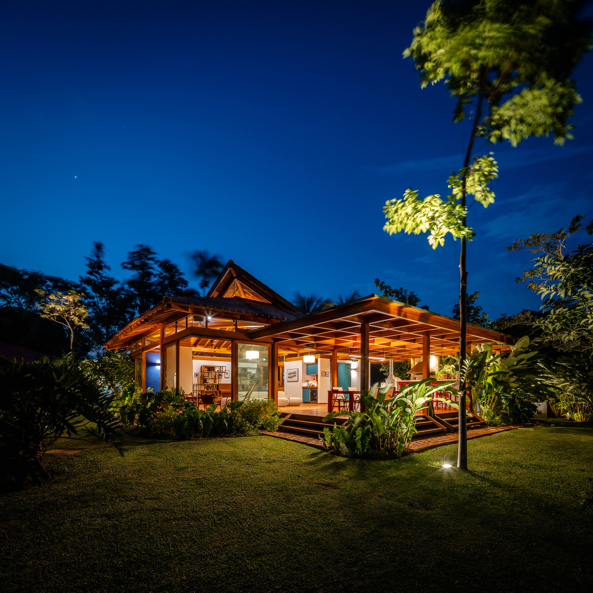 Single-story pavilion with pyramidal roof and timber pergola viewed across lawn under evening sky