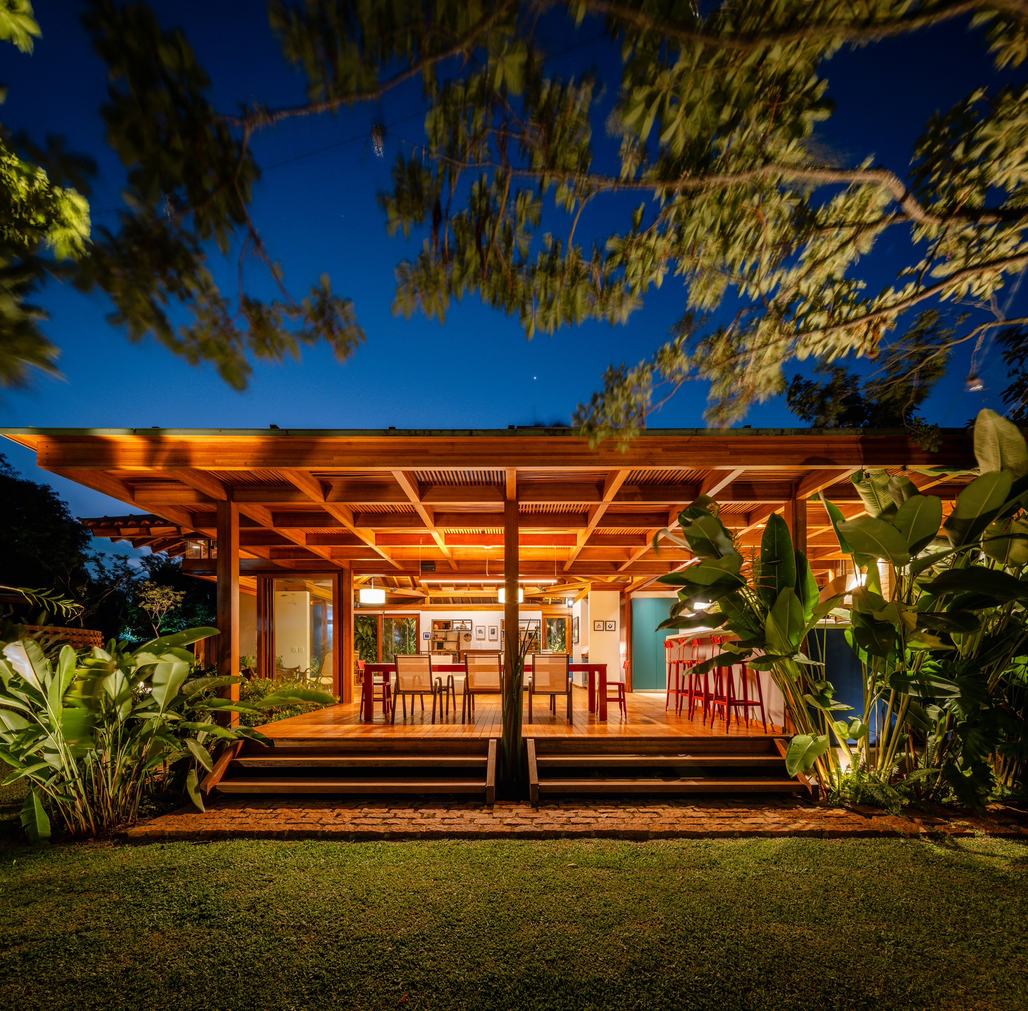 Flat timber pergola with slatted ceiling panels sheltering dining terrace and lawn at blue hour