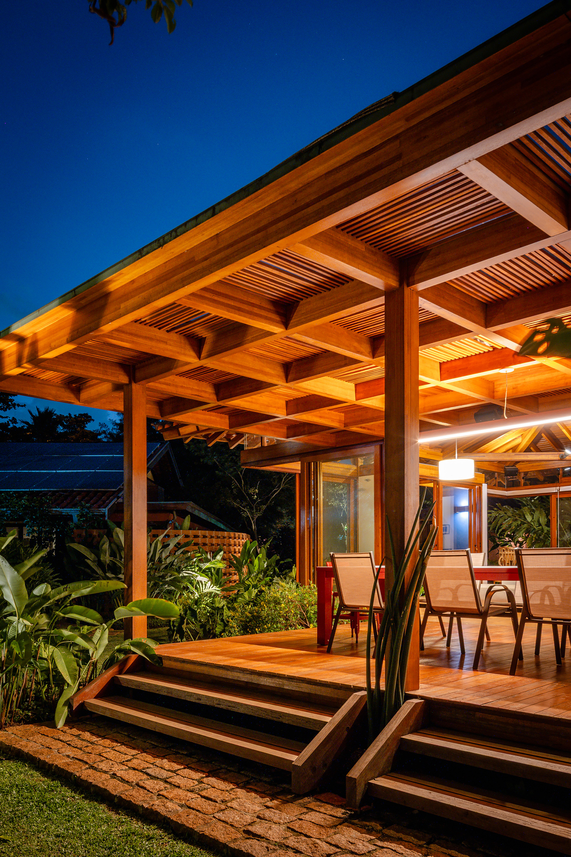 Detail of layered timber roof structure and stepped deck with chairs under illuminated eave