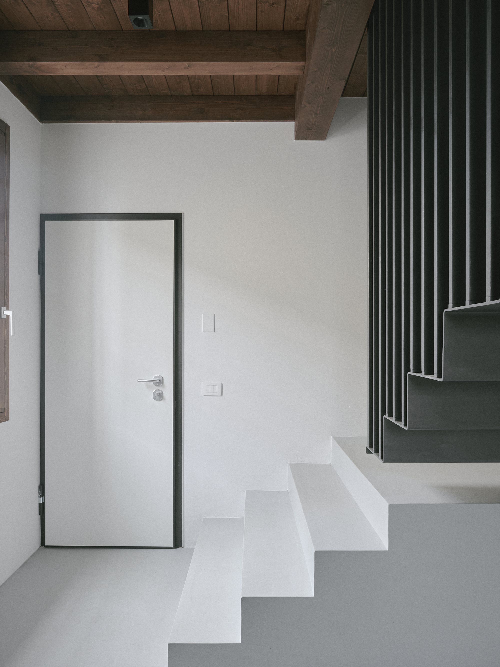 Entryway with white door beneath exposed timber beams and a staircase with metal balustrade
