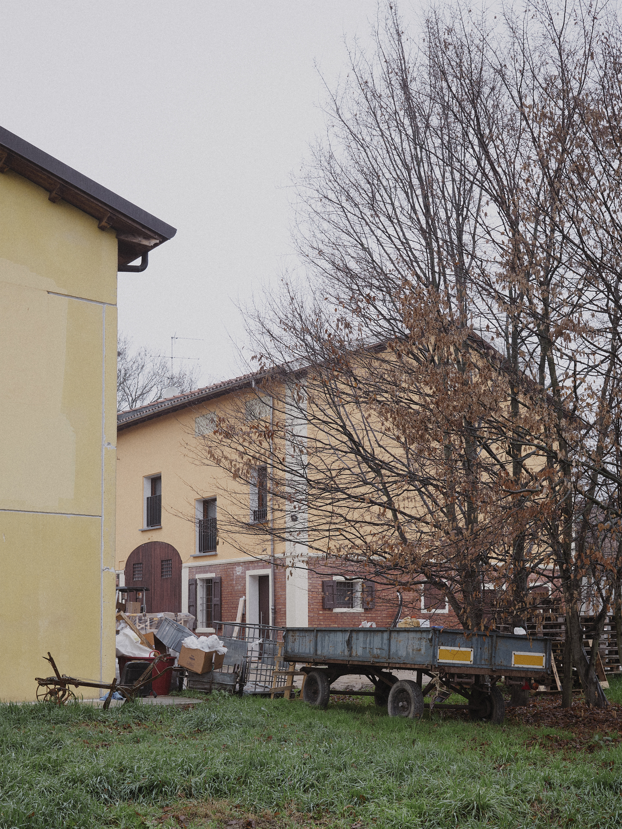 Yellow-rendered building volumes with bare deciduous trees and farm trailer in overcast weather
