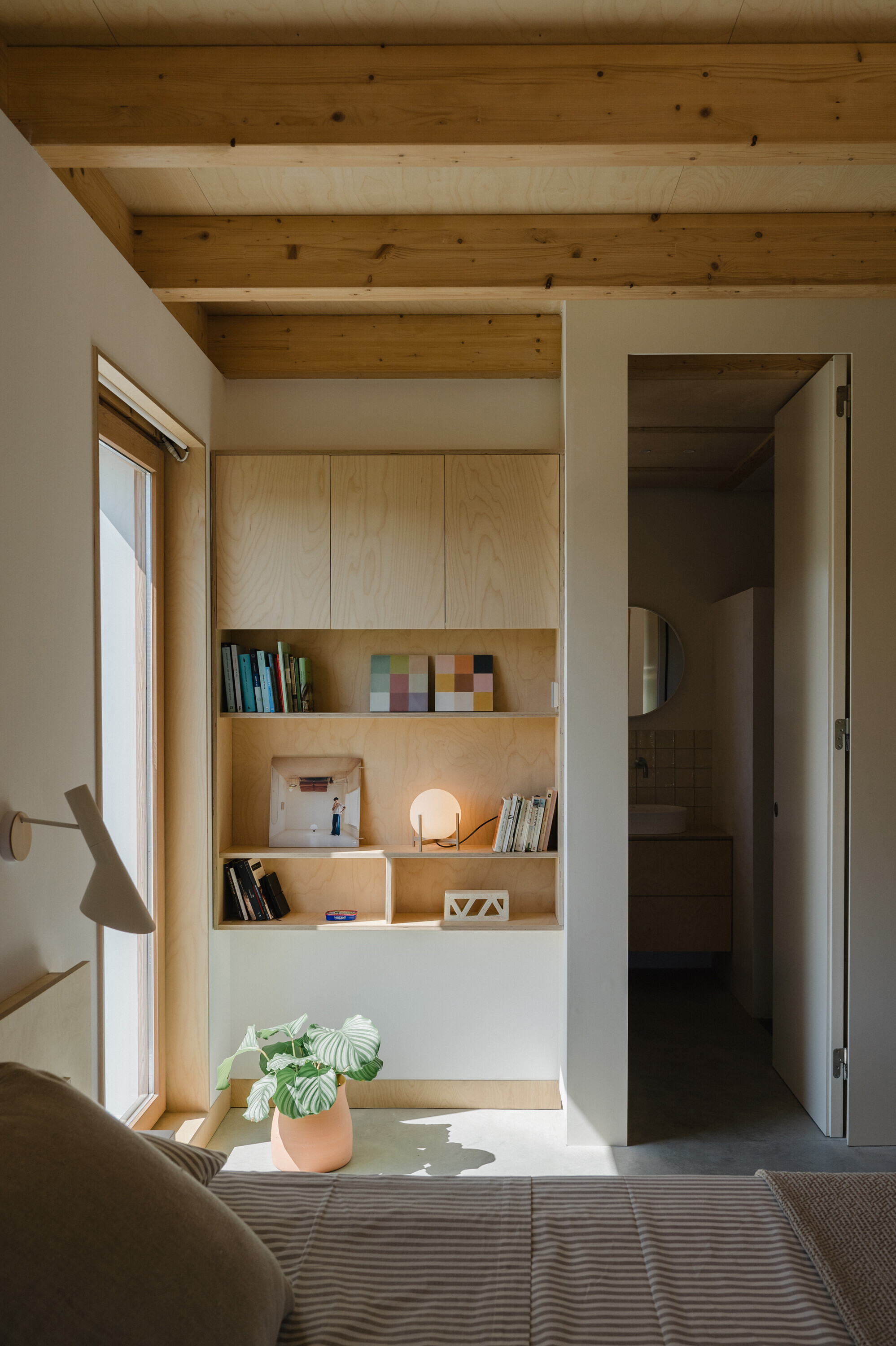 Bedroom with built-in plywood shelving niche beside doorway opening and window casting afternoon light
