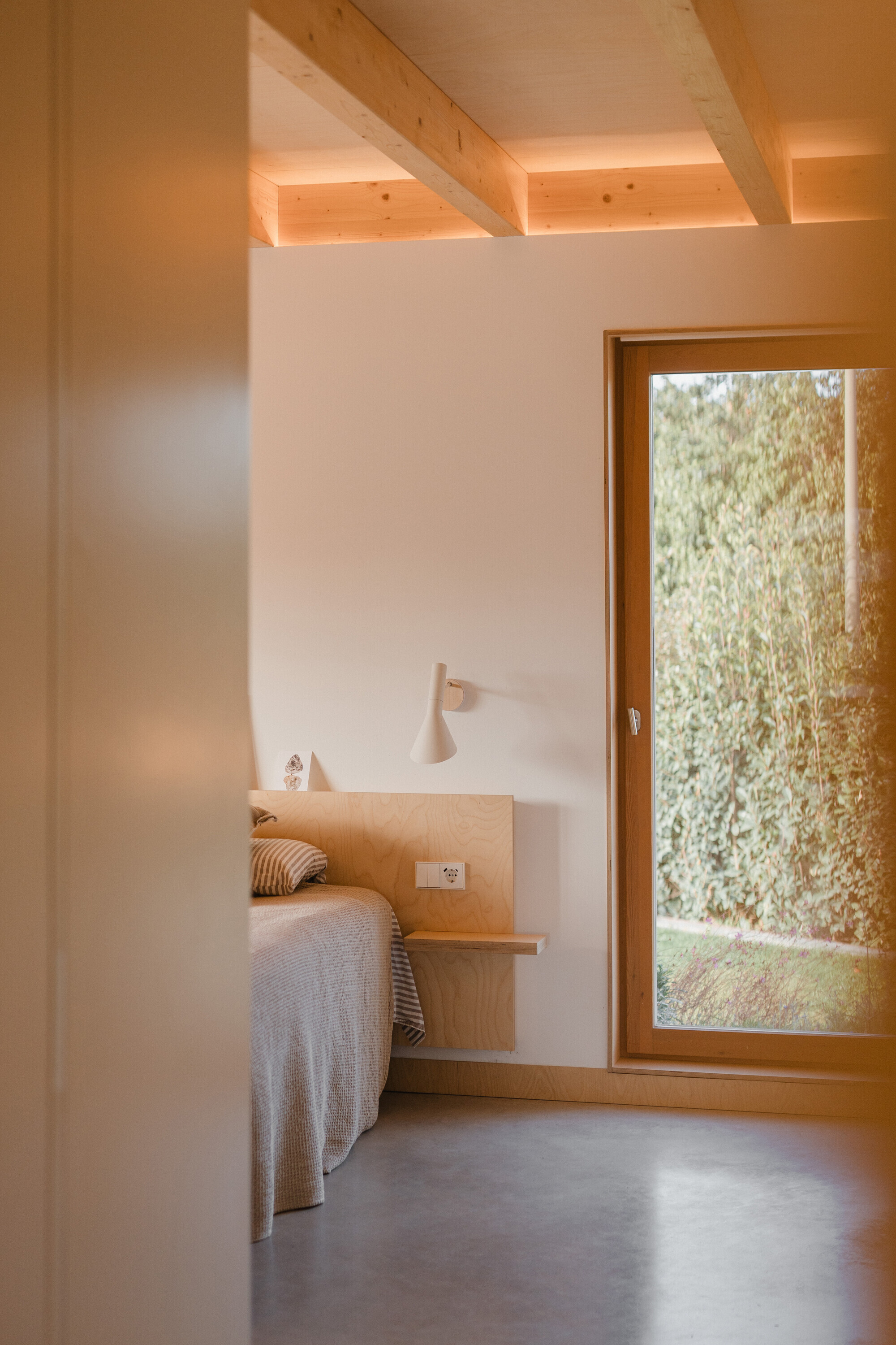 Bedroom with plywood bed frame and timber-beamed ceiling lit by afternoon sunlight through a garden-facing window