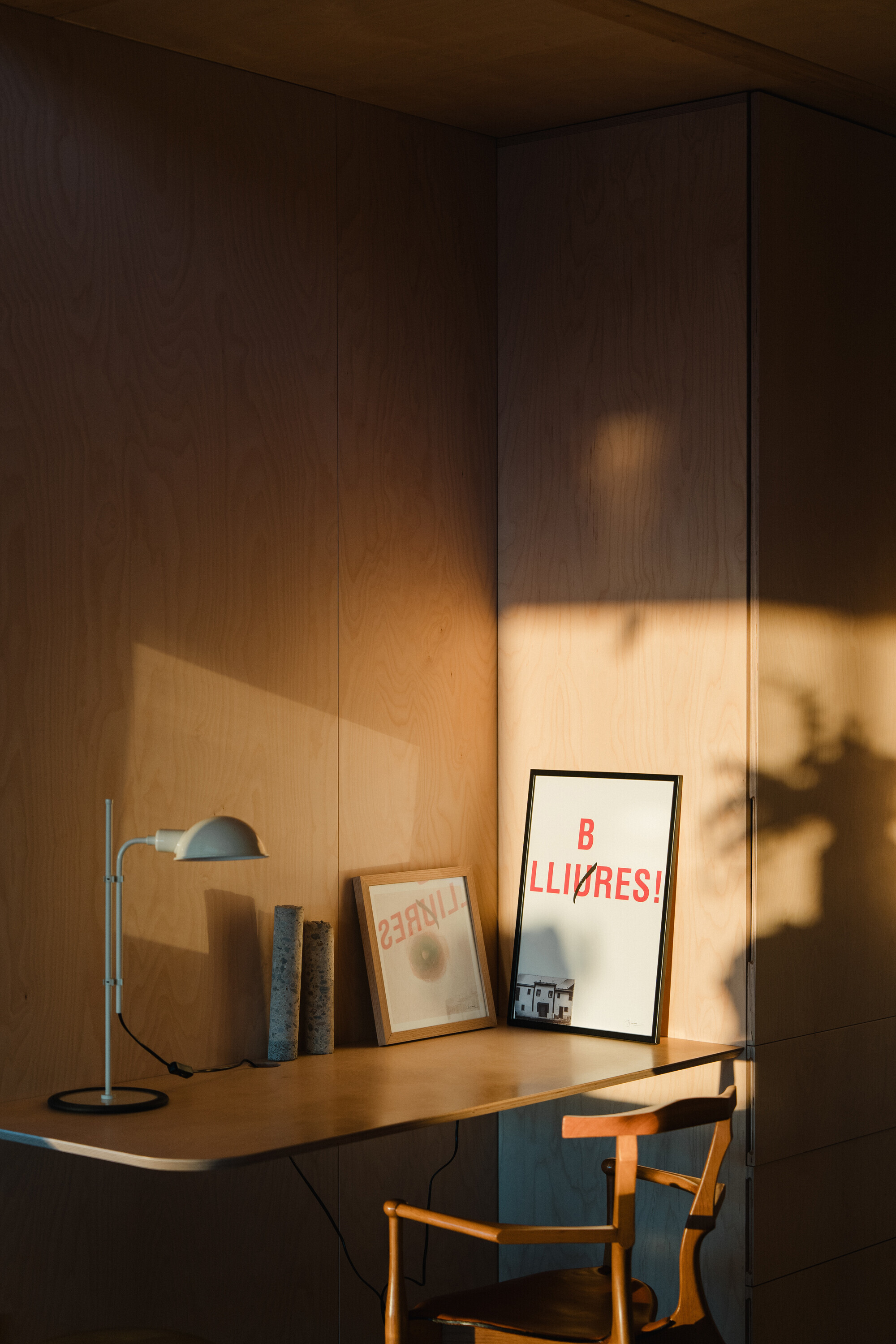 Plywood desk alcove with framed prints and desk lamp illuminated by golden evening light