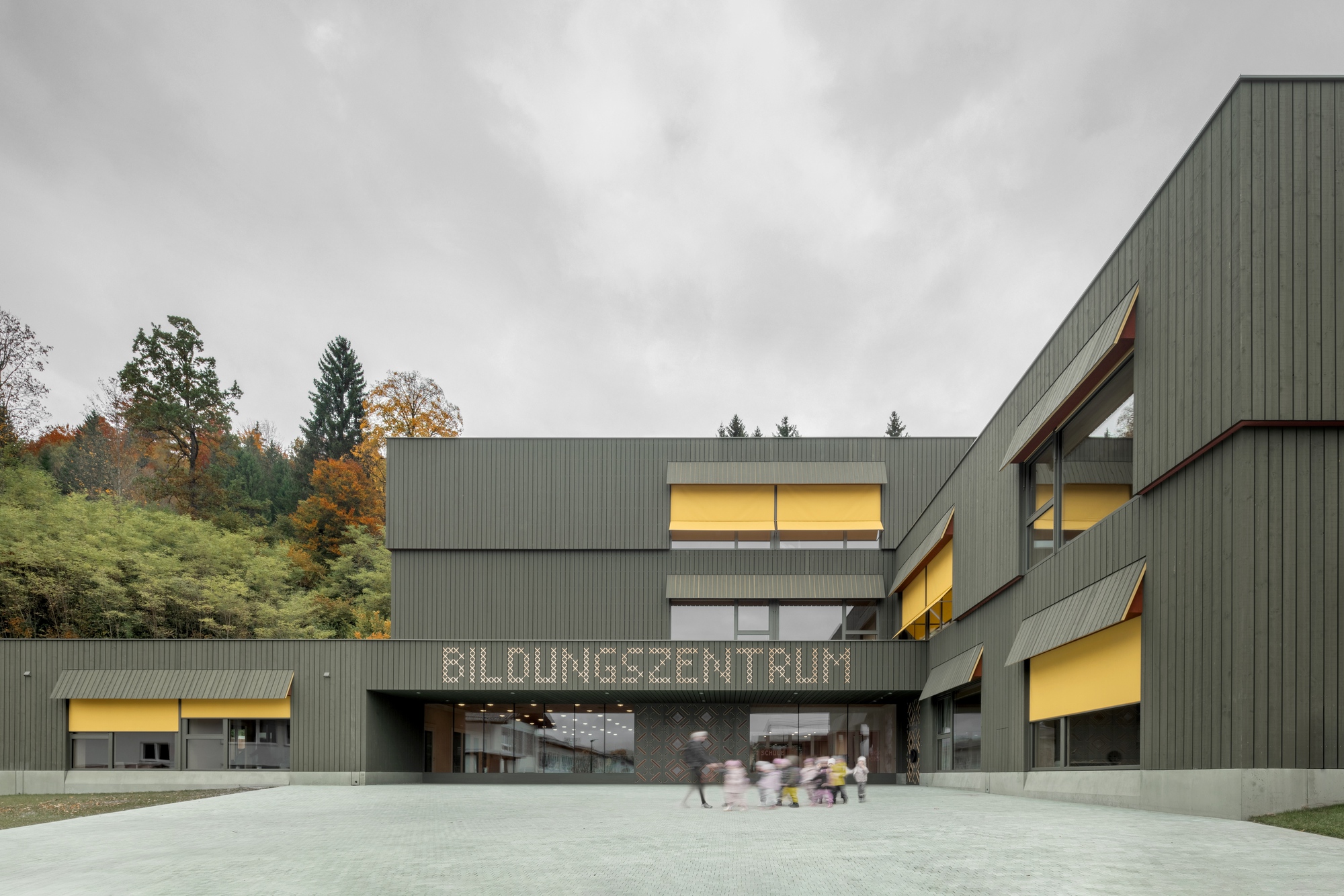 Corrugated metal facade with illuminated windows facing a concrete plaza at dusk beneath autumn trees