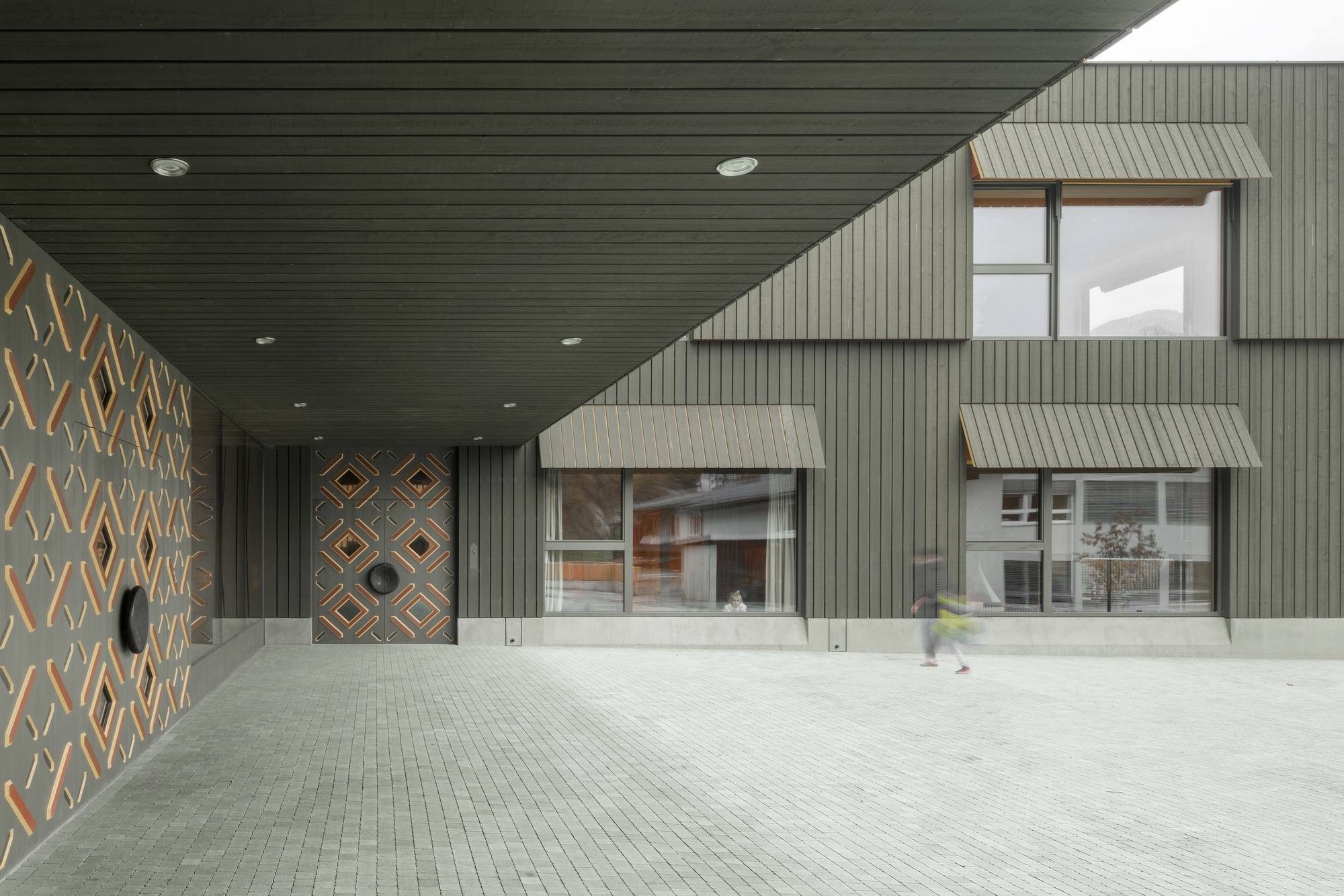 Covered entrance courtyard with timber-paneled ceiling and patterned wall adjacent to metal-clad facade volumes