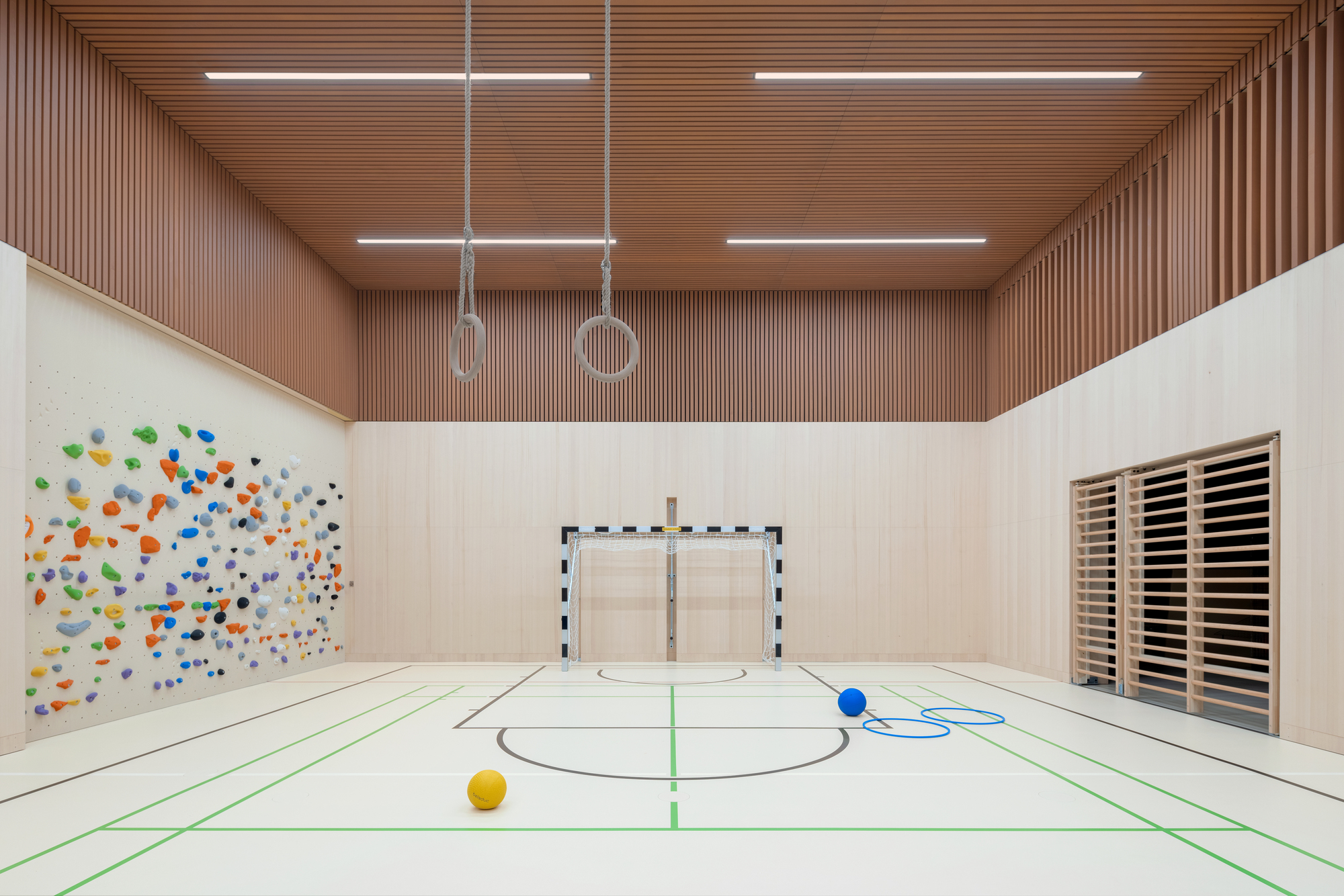 Indoor gymnasium with ribbed terracotta ceiling, floor markings, gymnastic rings, and a goal net against pale plywood walls