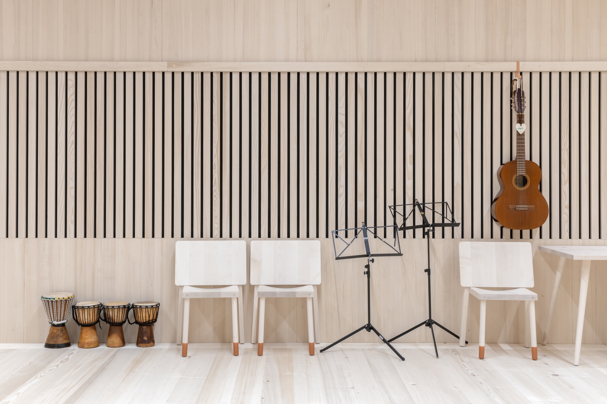 Music area with vertical slat wall, pale wood chairs, music stands, hand drums, and a guitar hanging overhead