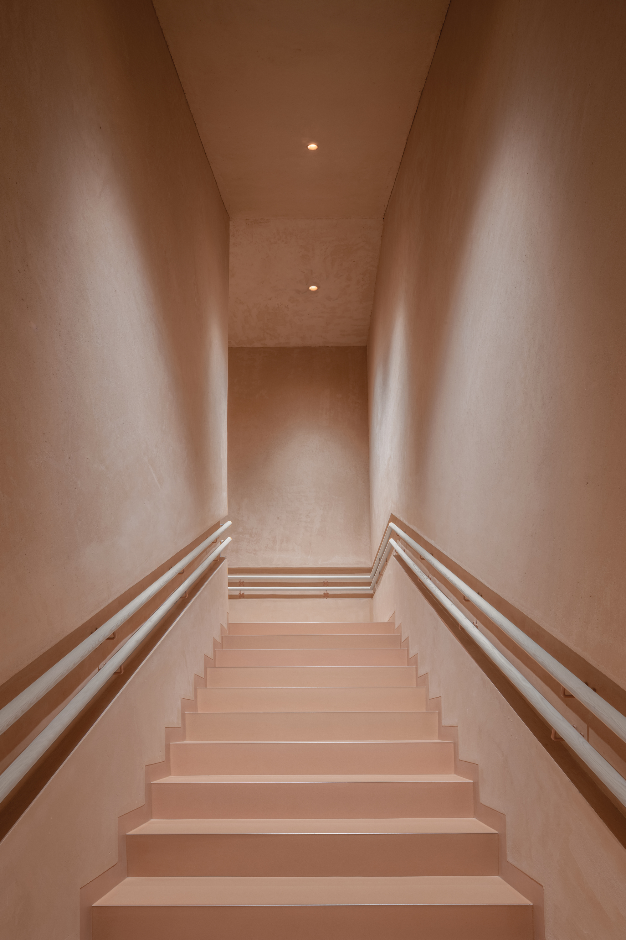 Narrow stairwell with timber handrails and pink tinted plaster walls ascending toward recessed ceiling lights
