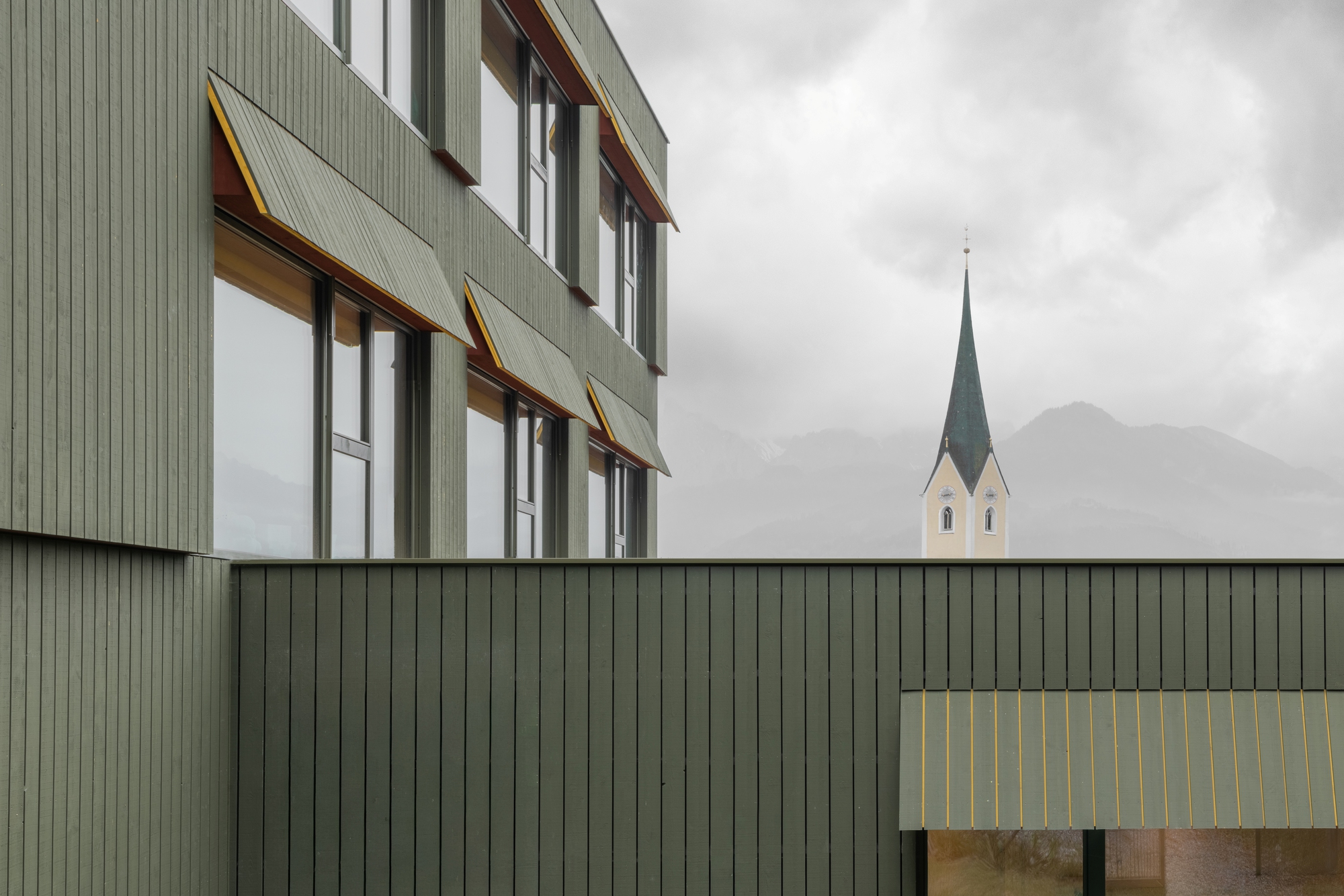 Green timber clad facade with angled window awnings and a distant church spire under overcast skies