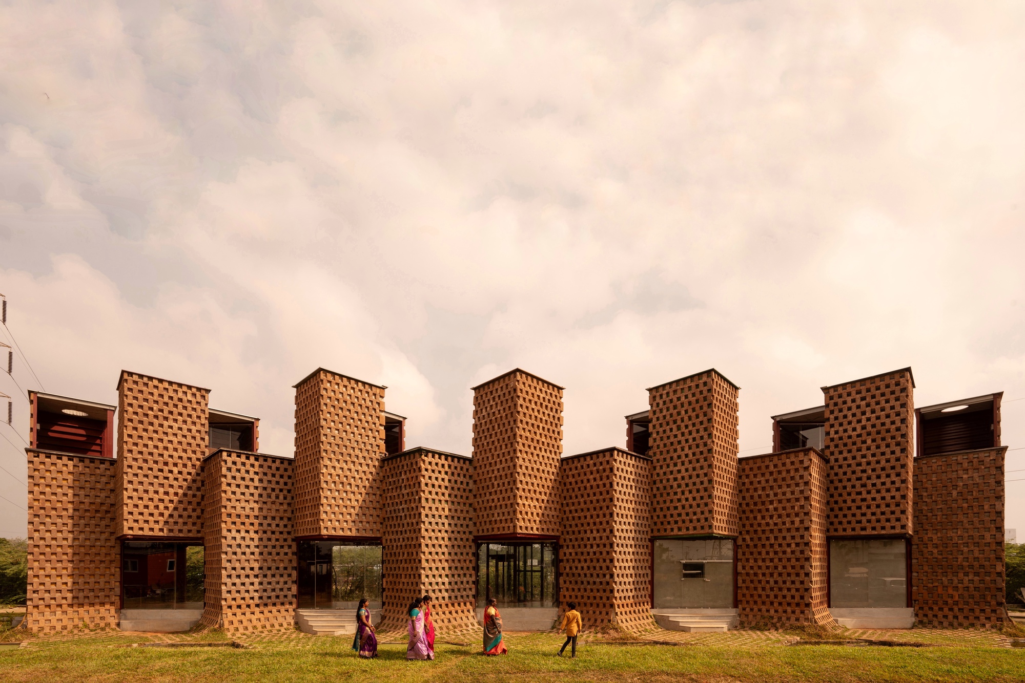 Perforated brick facade with crenellated towers and deep voids as people walk across the lawn