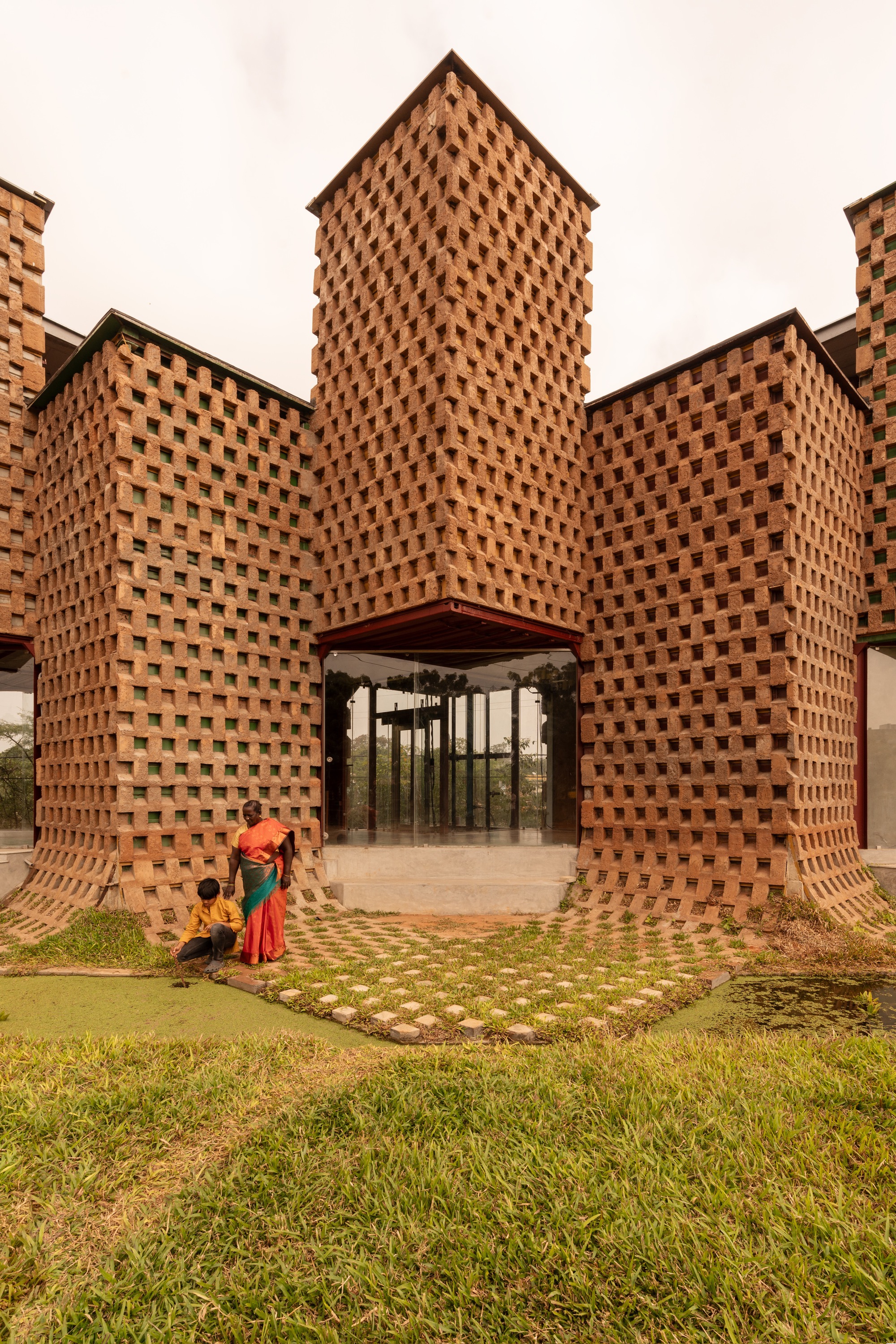 Central arched entrance through perforated brick towers framed by people and a grass paver courtyard