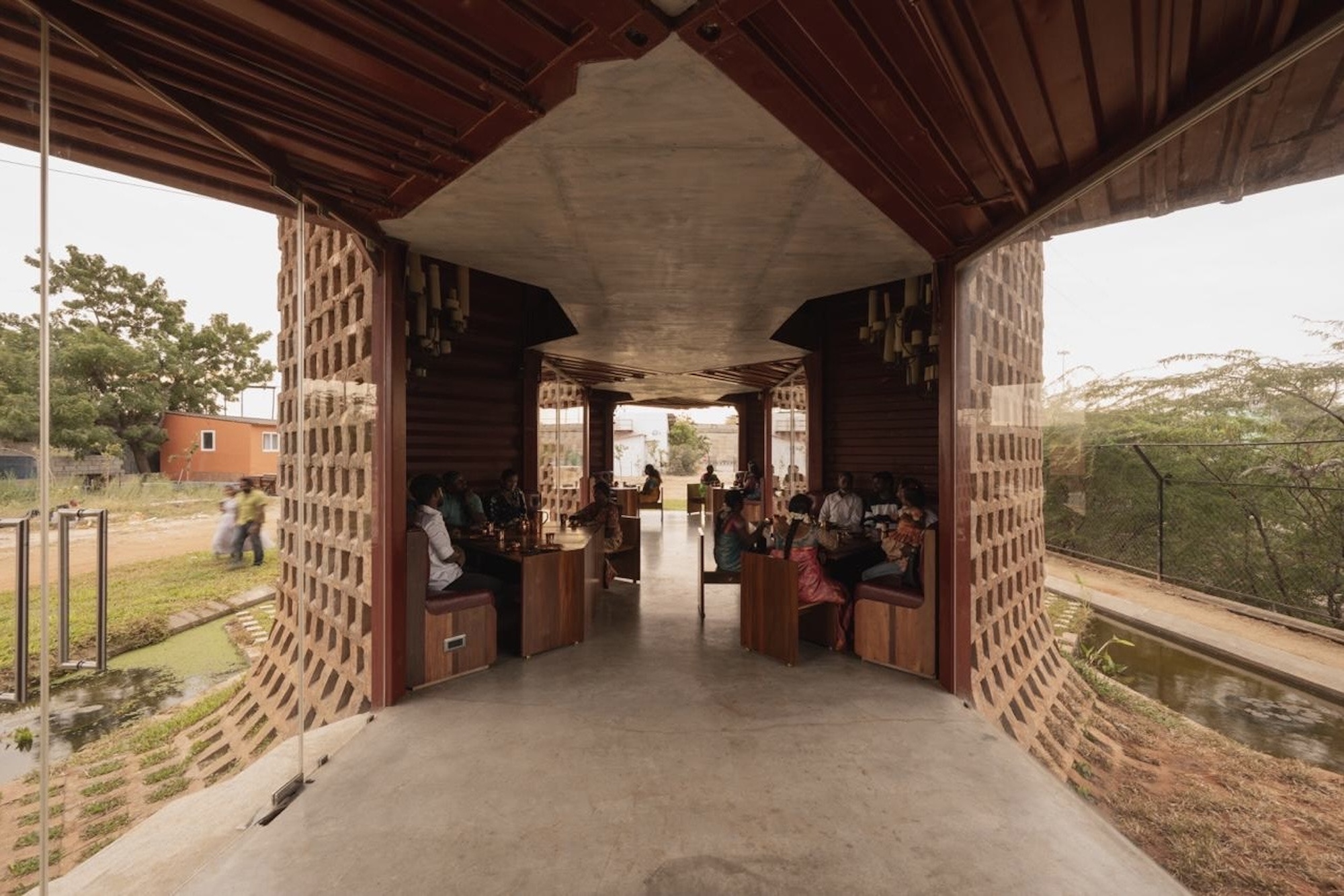 Covered passage with timber ceiling panels and concrete beams between perforated brick walls with seated visitors