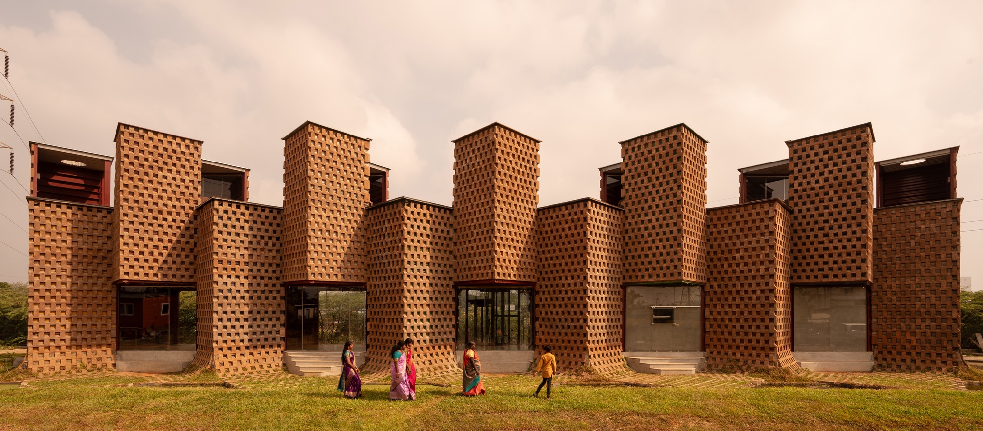 Perforated brick facade with cylindrical columns and glass openings as people walk across the lawn