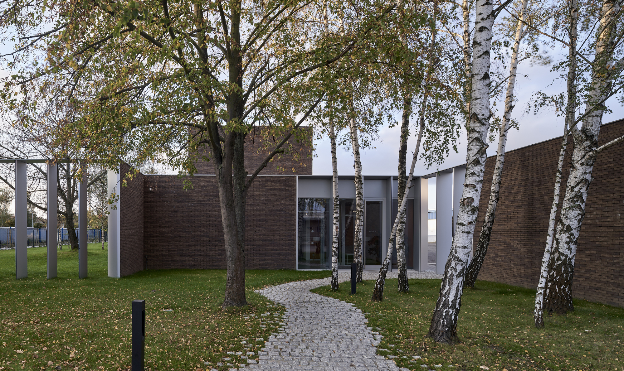 Cobblestone pathway leading through birch trees toward a courtyard entrance between brick volumes