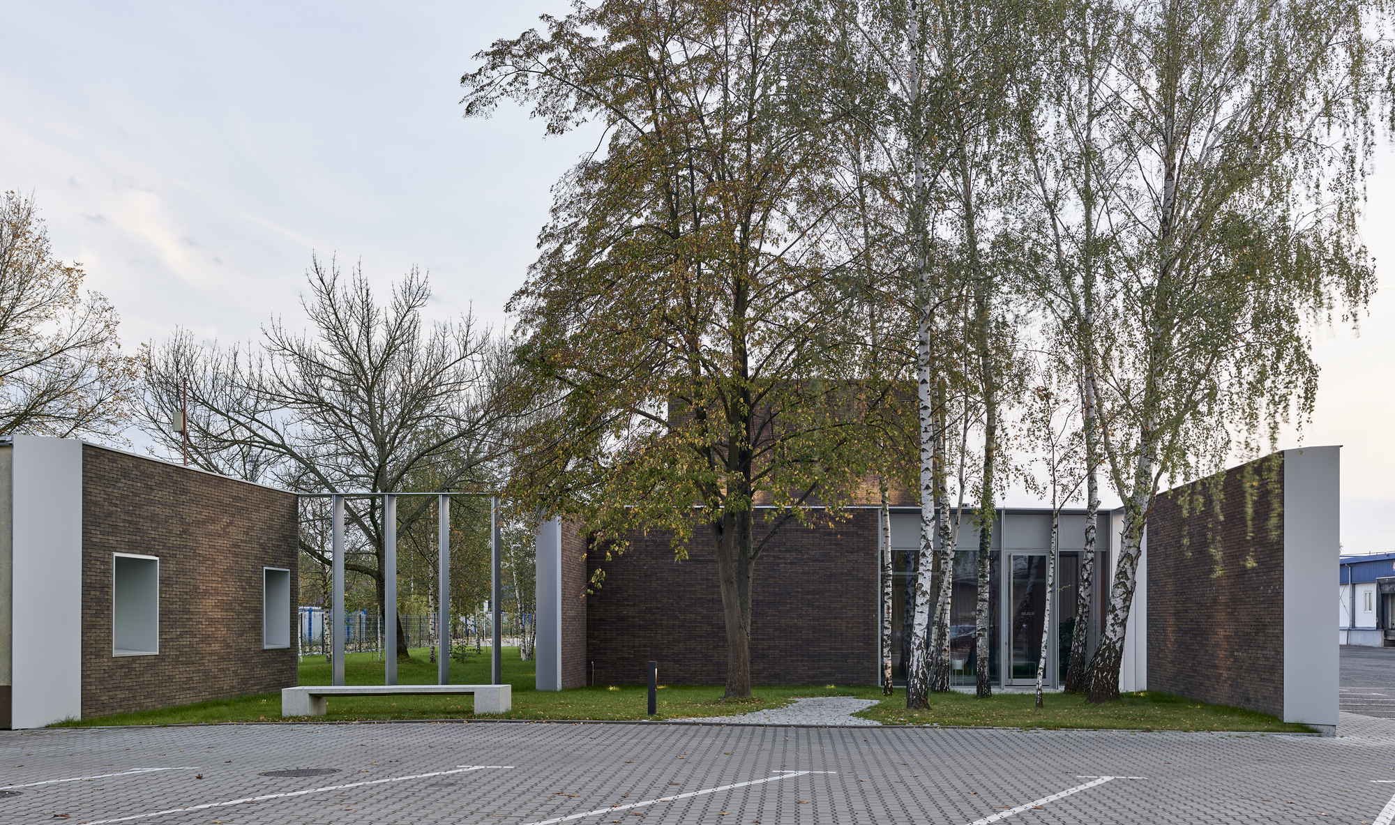 Street view of the dark brick and white panel volumes with birch trees and benches in a courtyard setting