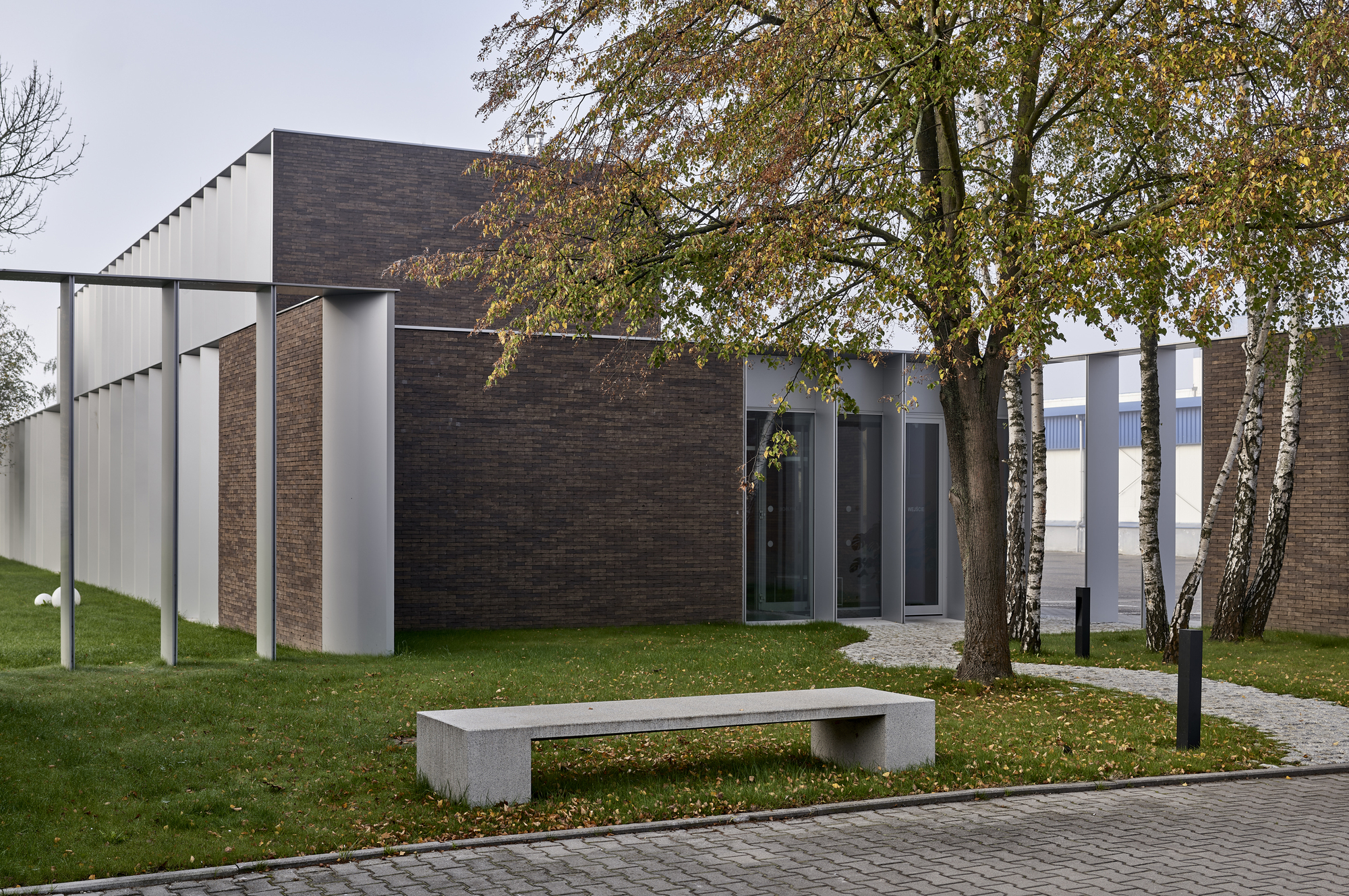 Dark brick facade with vertical metal and glass openings framed by birch trees on a grass lawn