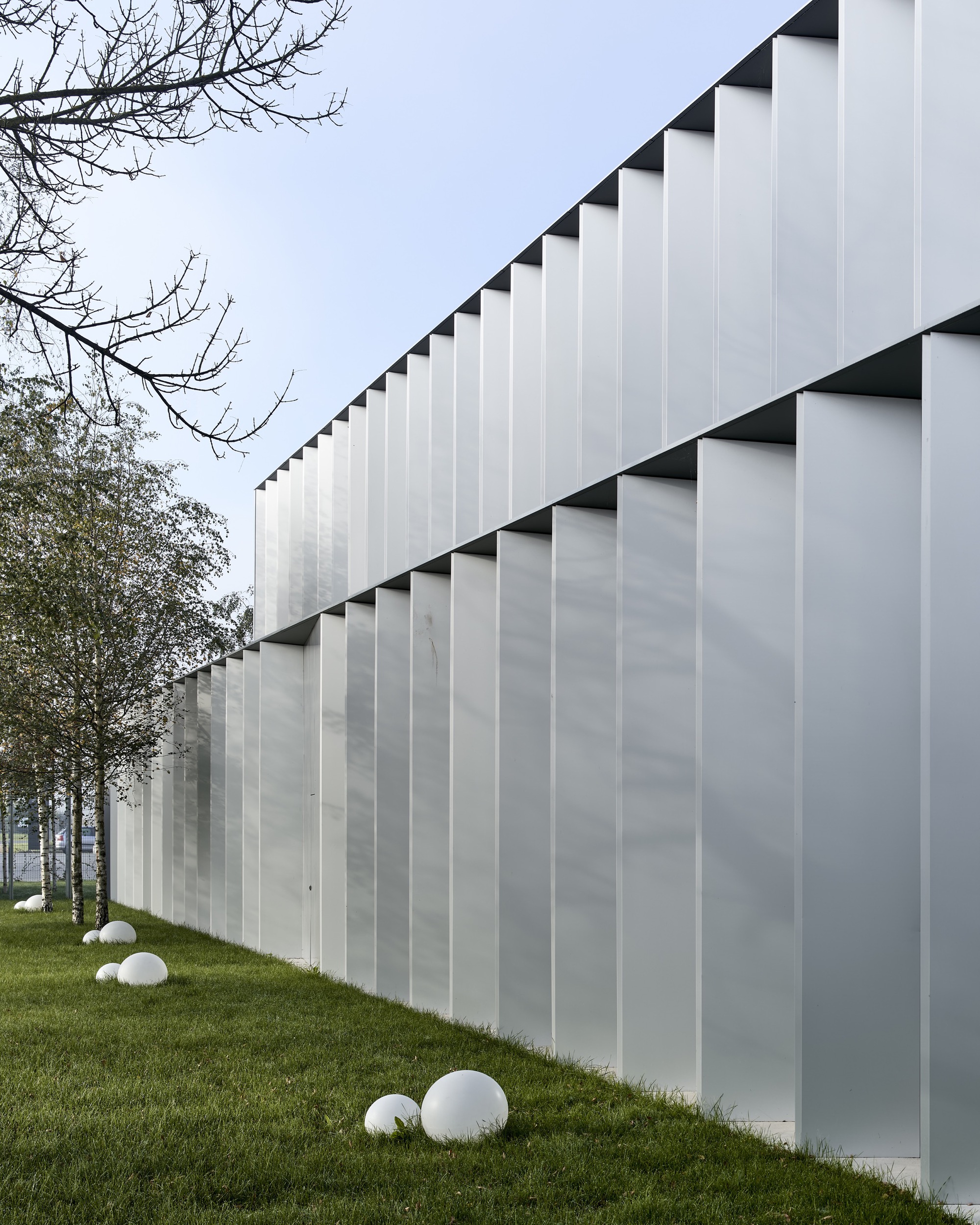 Translucent vertical panel facade with triangular shadow reveals beside a sloping lawn and bare trees