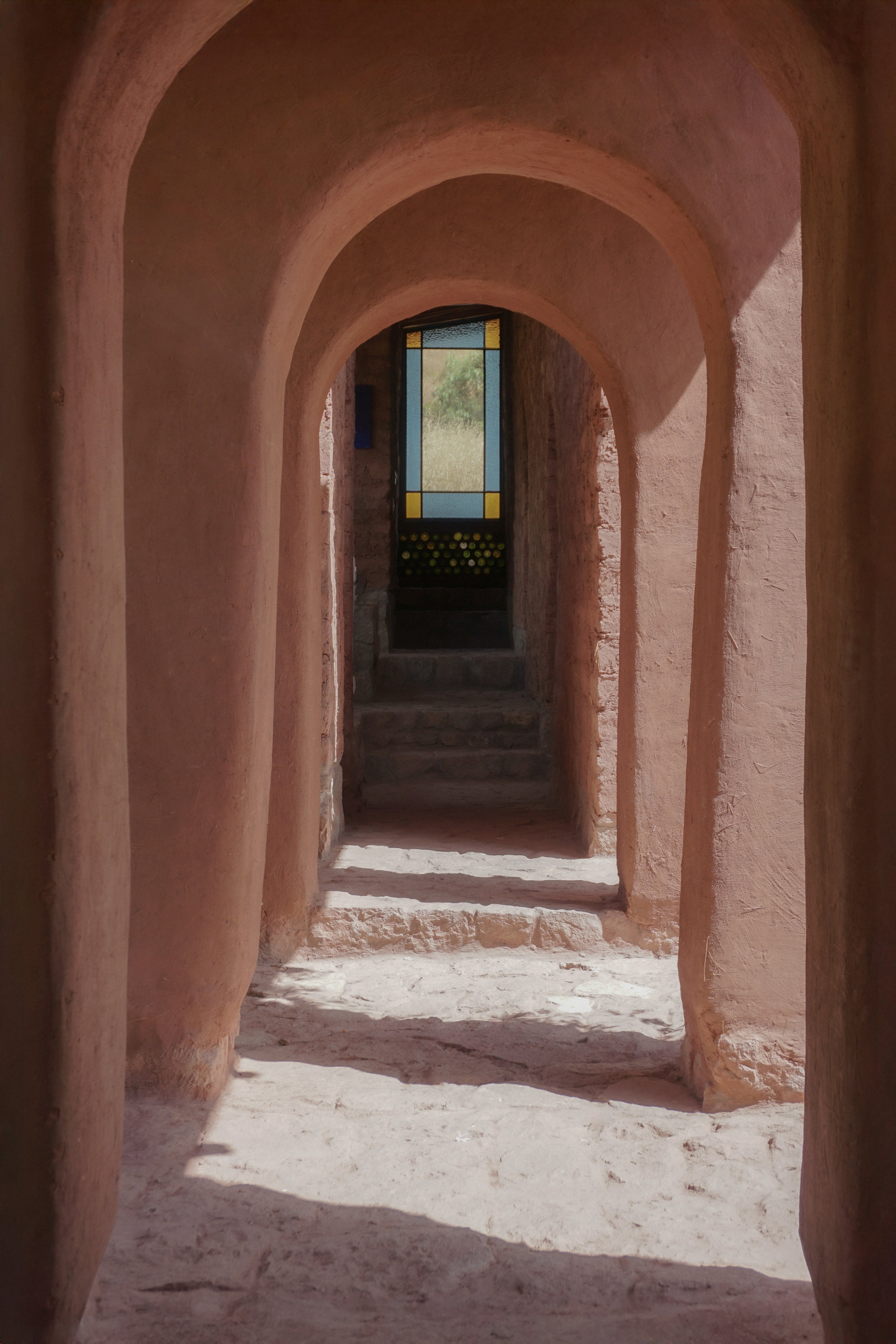 Sequence of rounded arches in earthen plaster corridor with stained glass door at far end