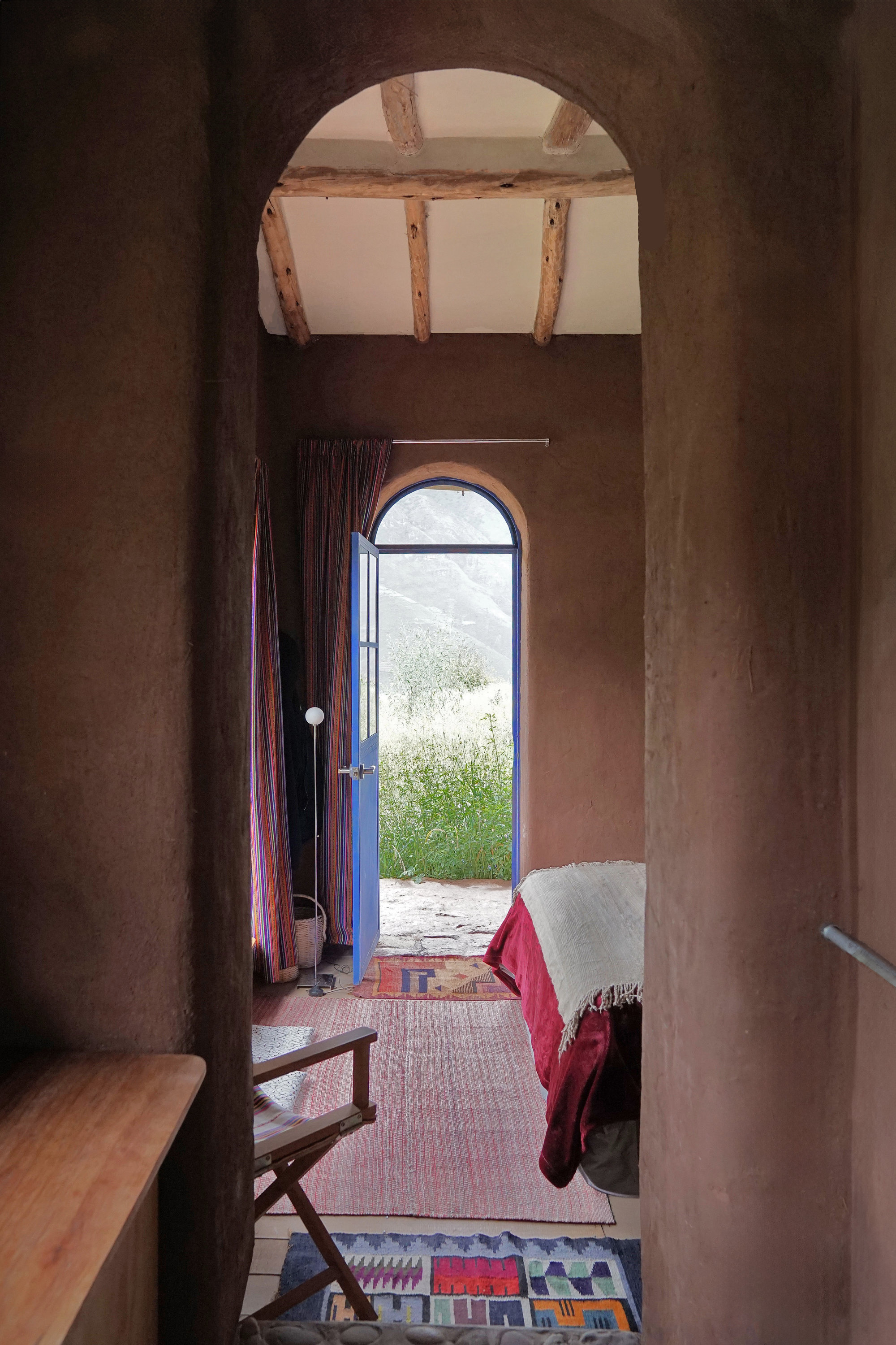 Arched doorway framing a bedroom with exposed timber ceiling beams and an open entrance to the landscape