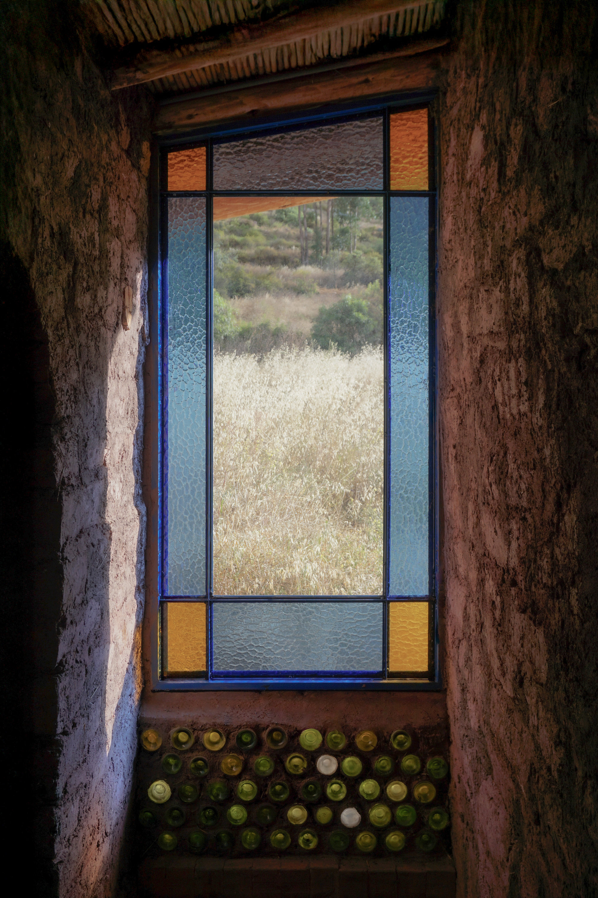 Tall window with colored glass panes and a grid of glass bottles embedded in the earthen sill