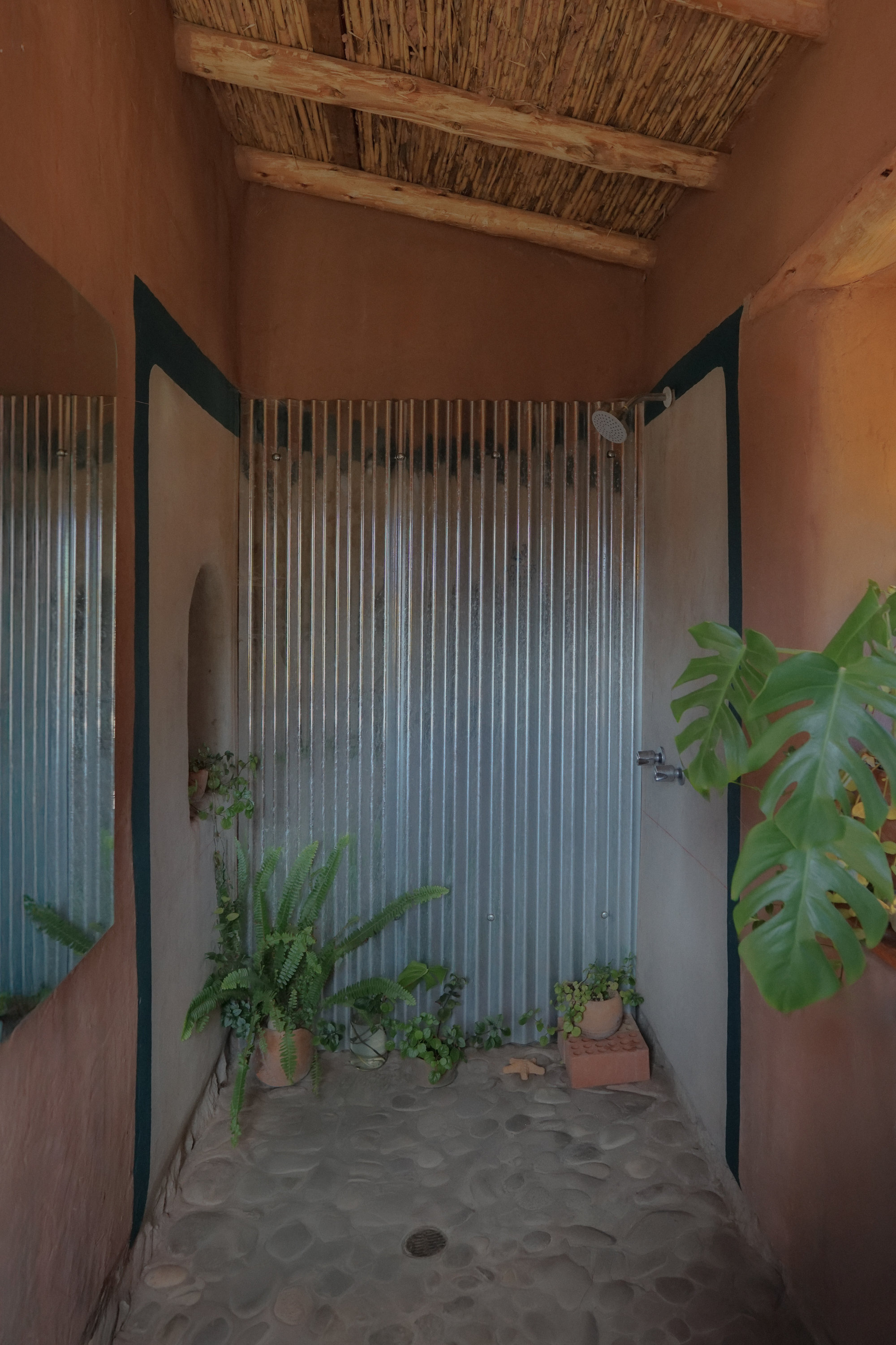 Outdoor shower enclosure with corrugated metal walls, reed ceiling and potted plants on stone paving