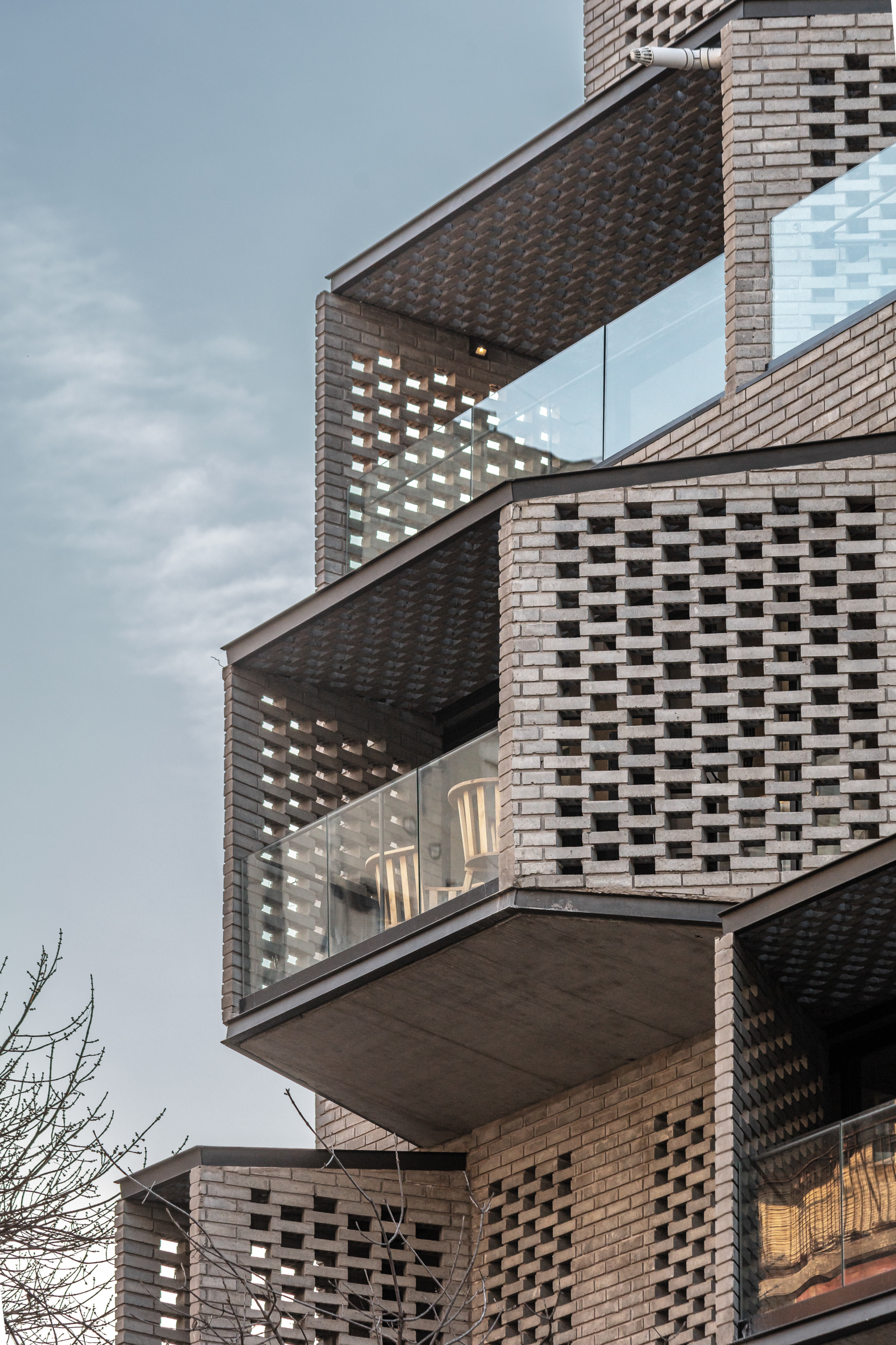 Upward view of cantilevered balconies clad in perforated brick with glass railings under overcast sky