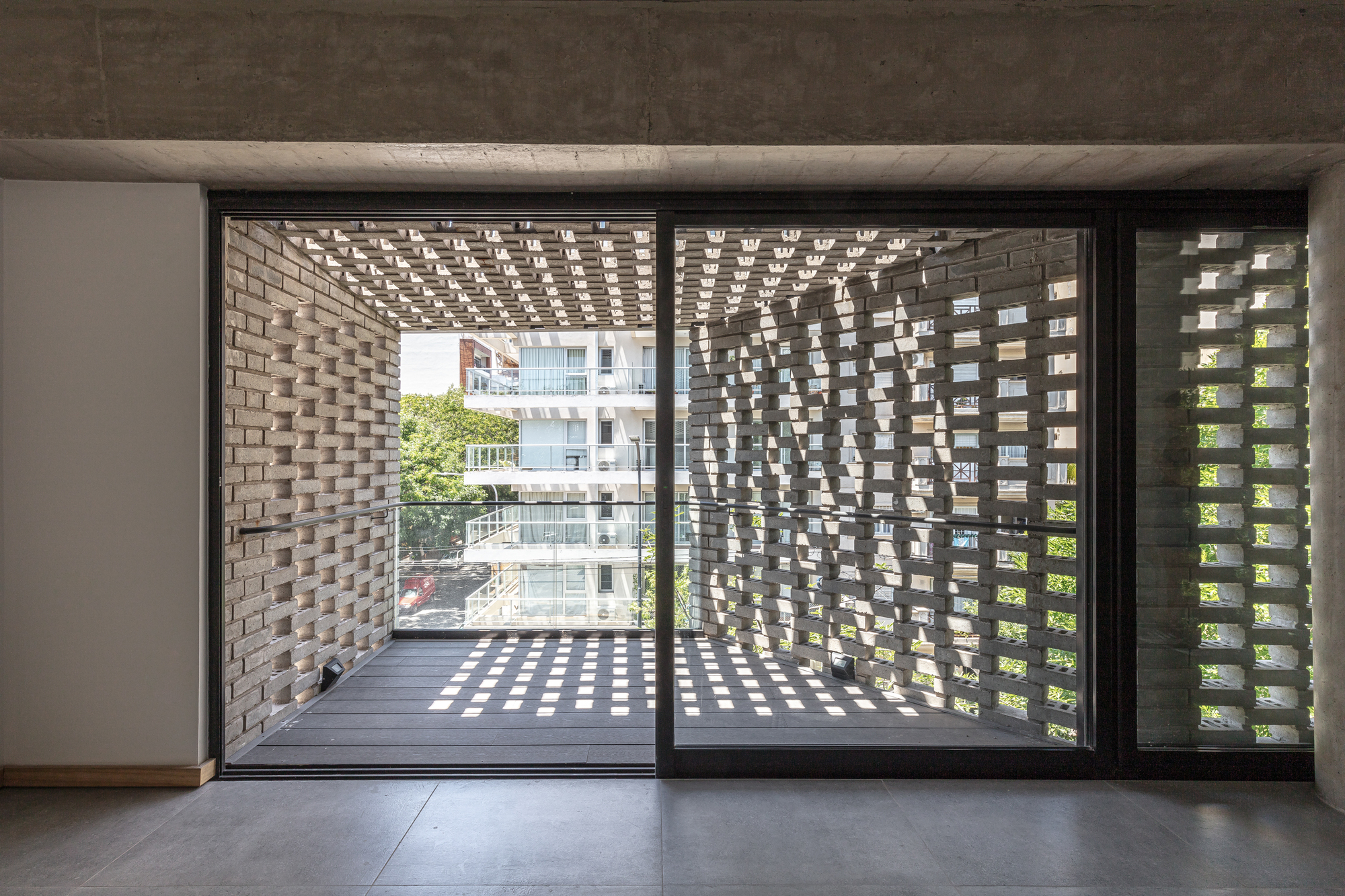 Interior view through sliding glass doors to a balcony enclosed by perforated brick screens casting dappled light