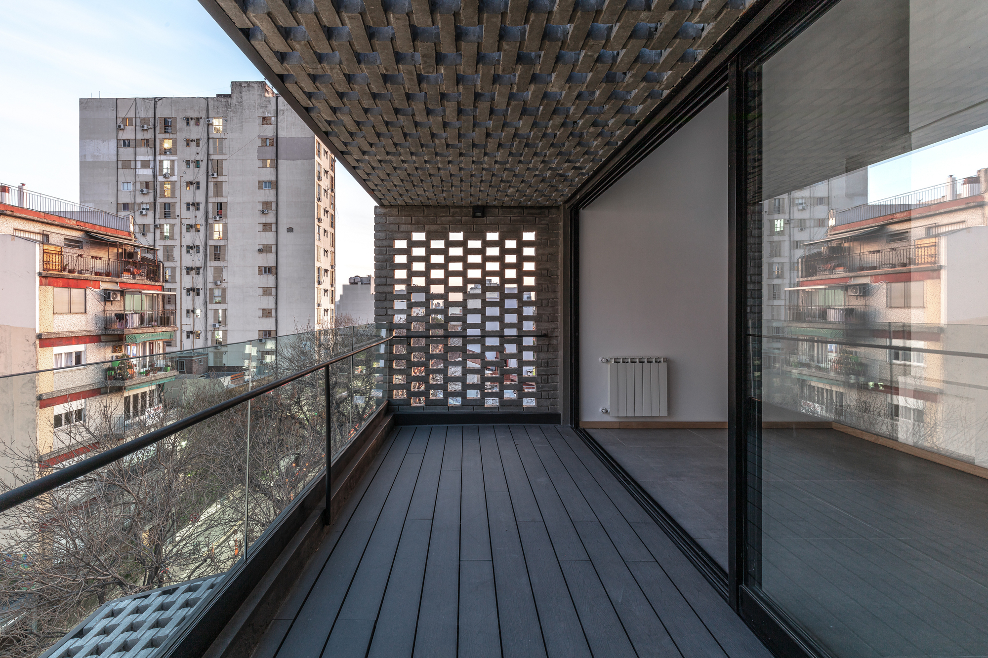 Exterior terrace with dark wood decking under a patterned brick soffit and glass railing at dusk