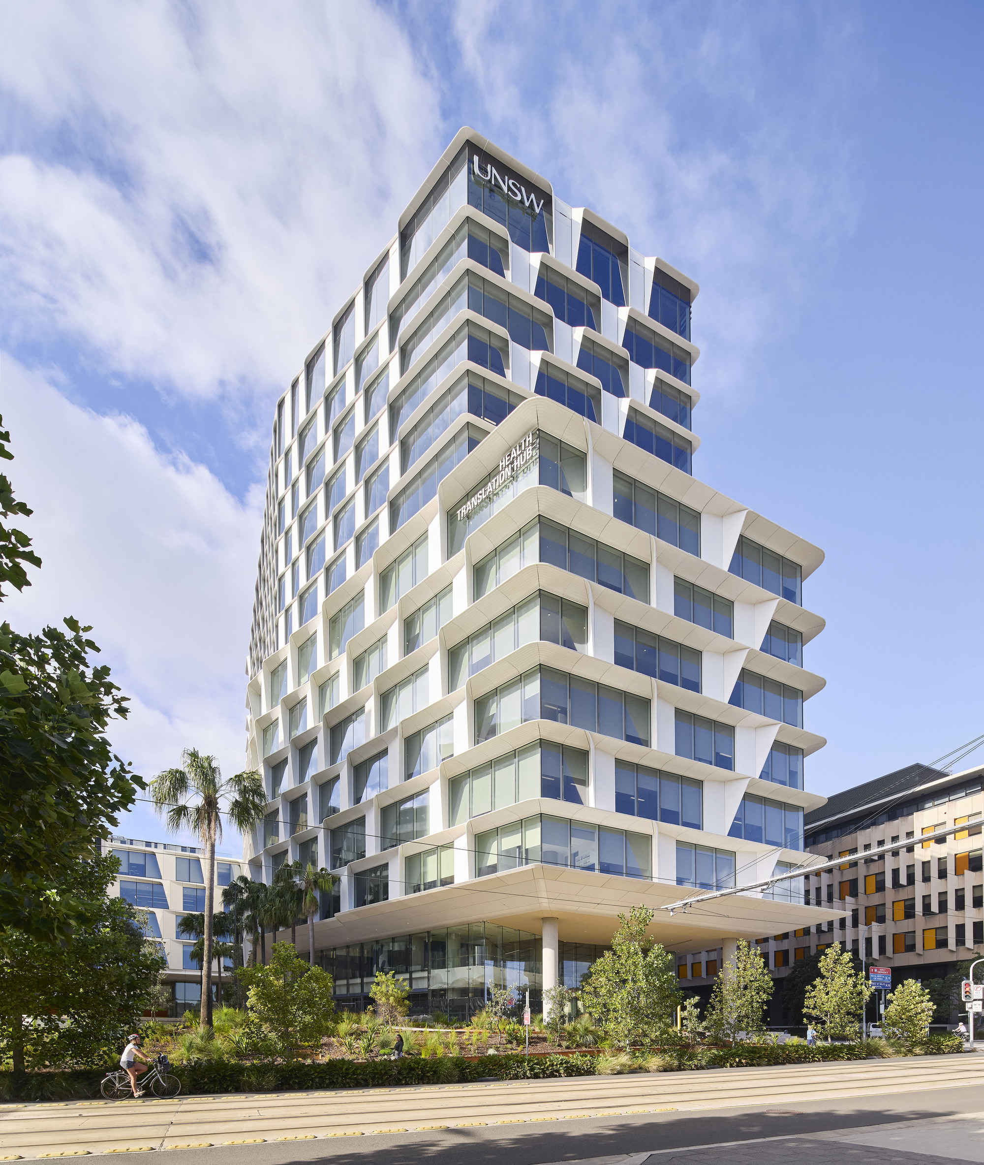 Street-level view of the cantilevered white facade with cyclist passing palm trees and planted beds