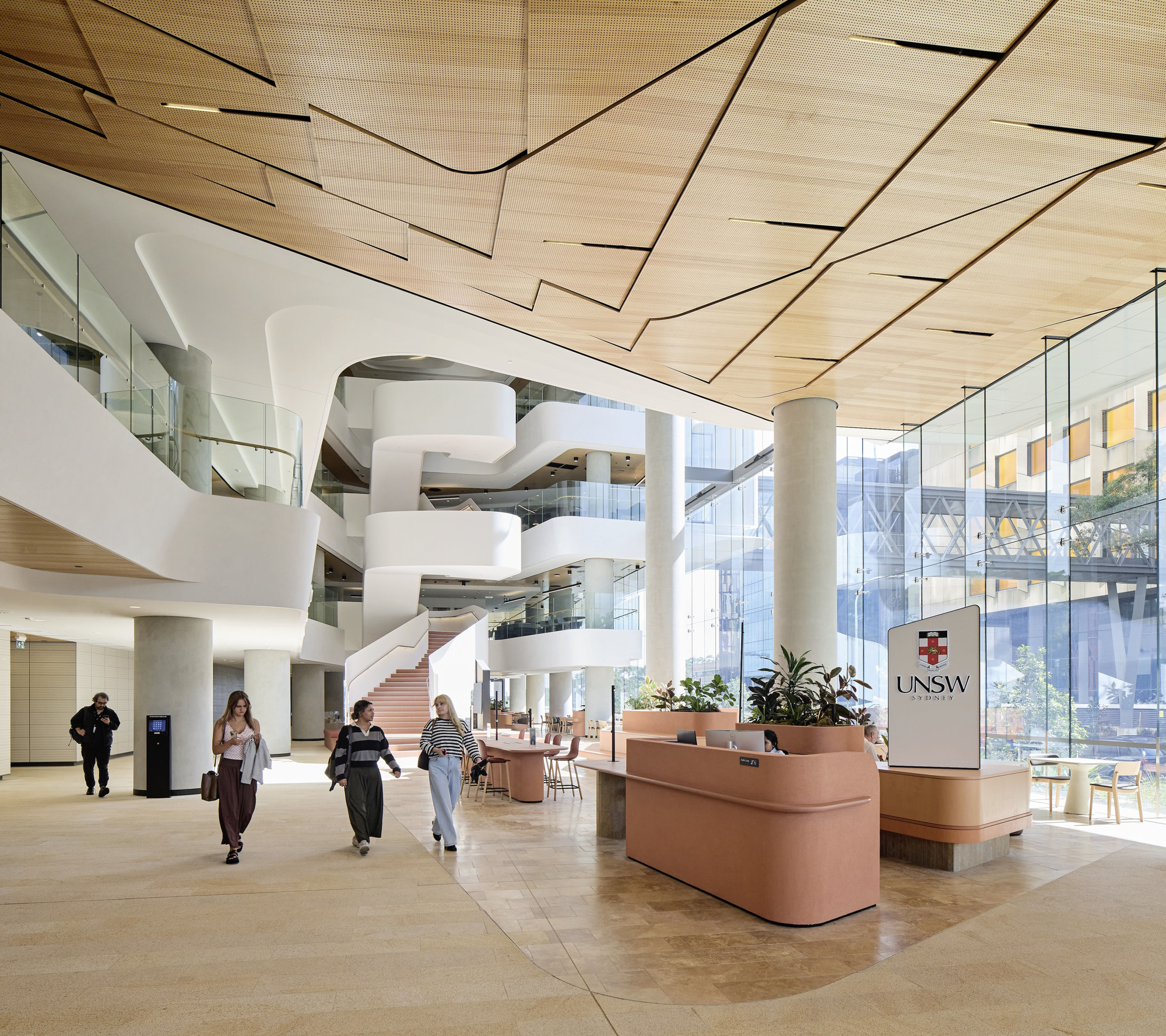 Atrium with terrazzo flooring, white curved balconies, timber ceiling planes and students entering beneath glazed facades