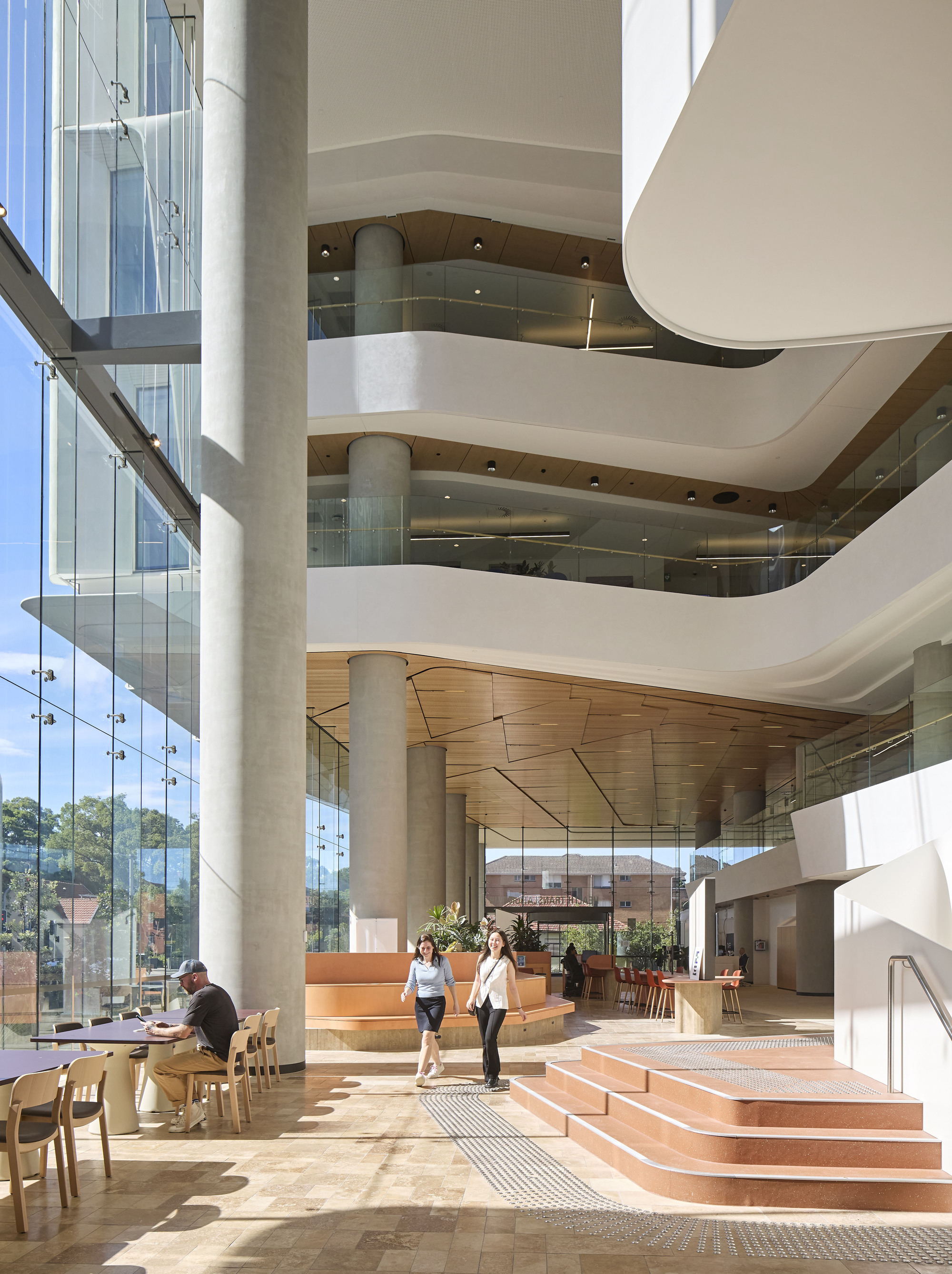 Stacked atrium balconies with curved white edges and timber soffits above terracotta-toned stepped seating