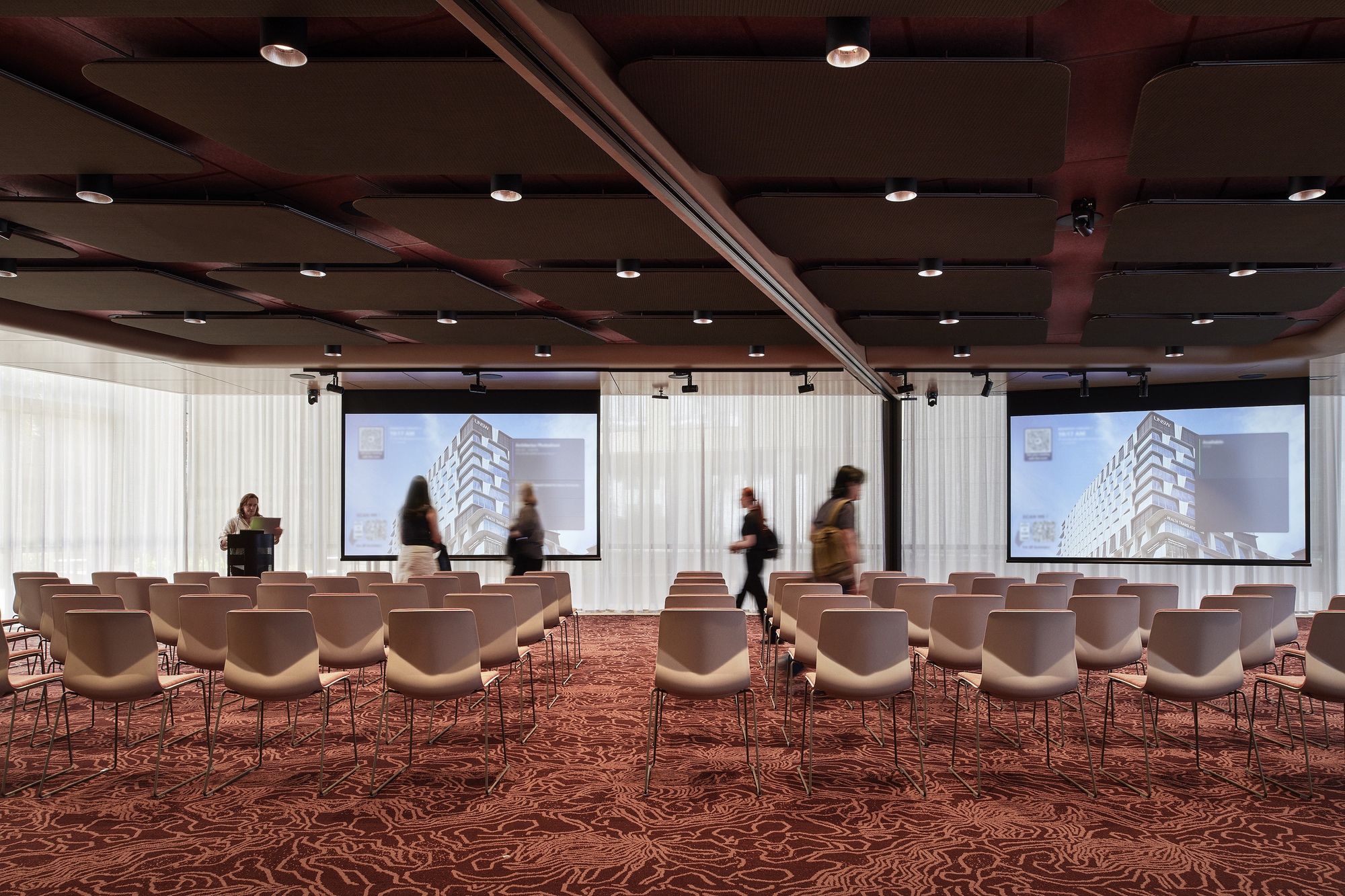Conference room with patterned burgundy carpet, white chairs and people moving past projection screens beneath dark timber ceiling