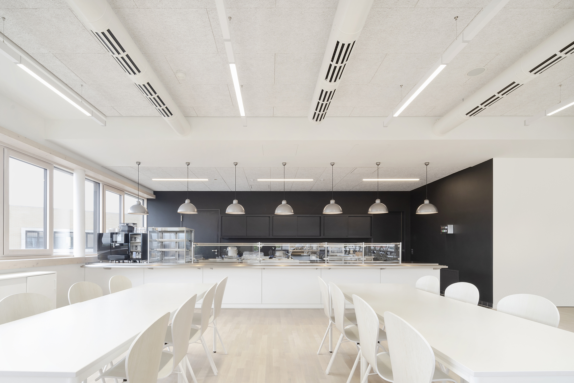 Communal kitchen with black wall-mounted cabinetry and pendant lights hung above white tables