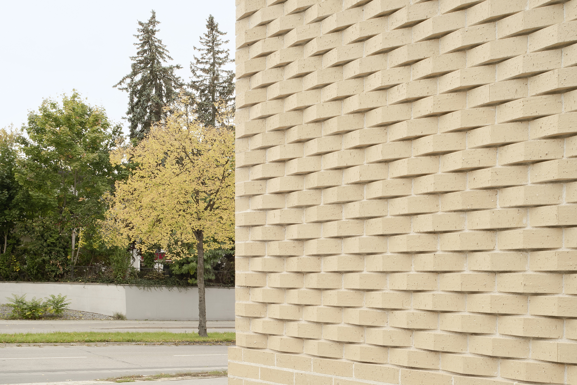Textured brick facade with projecting headers beside a young tree in autumn leaf