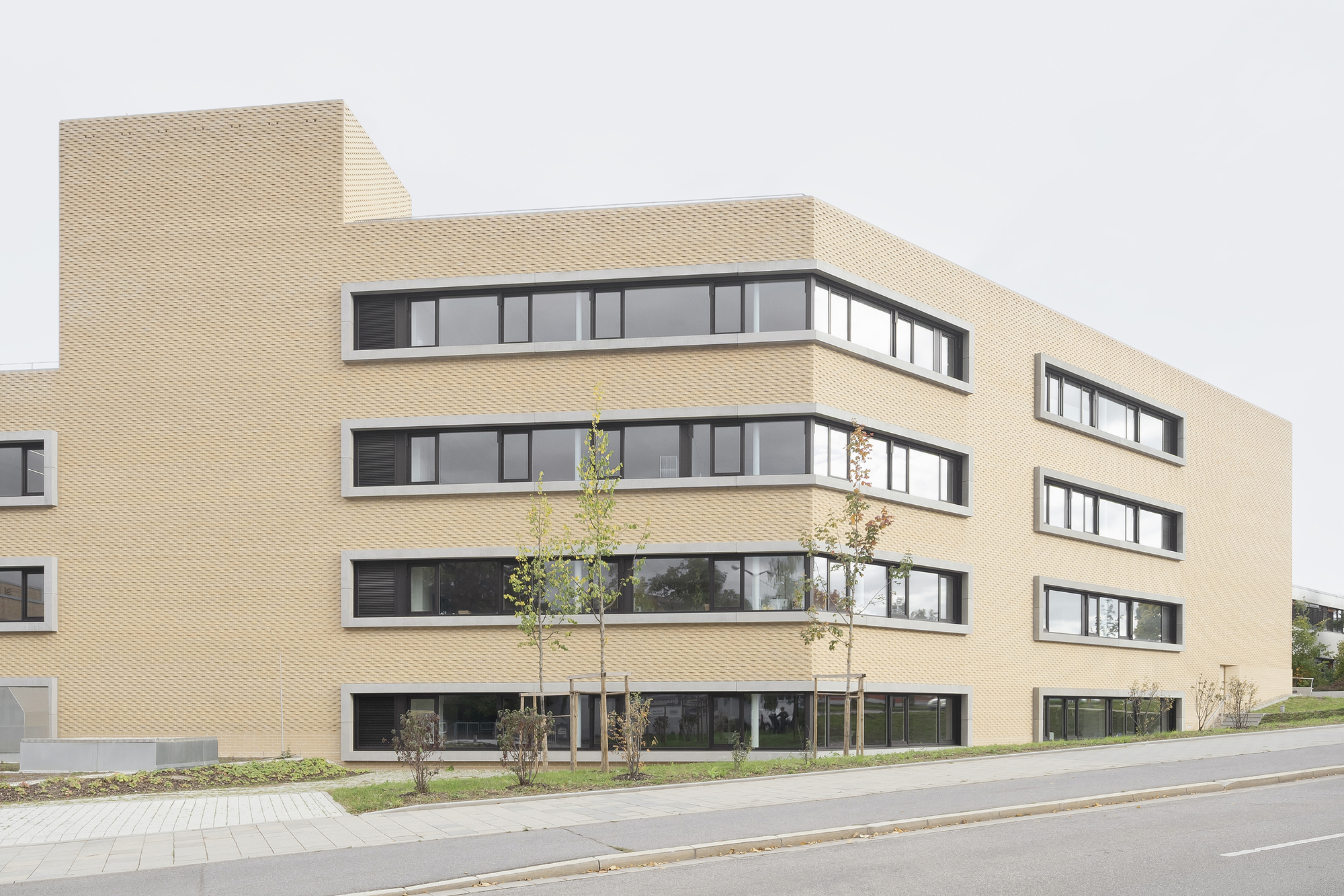 Street view of the textured brick facade with ribbon windows and recently planted saplings