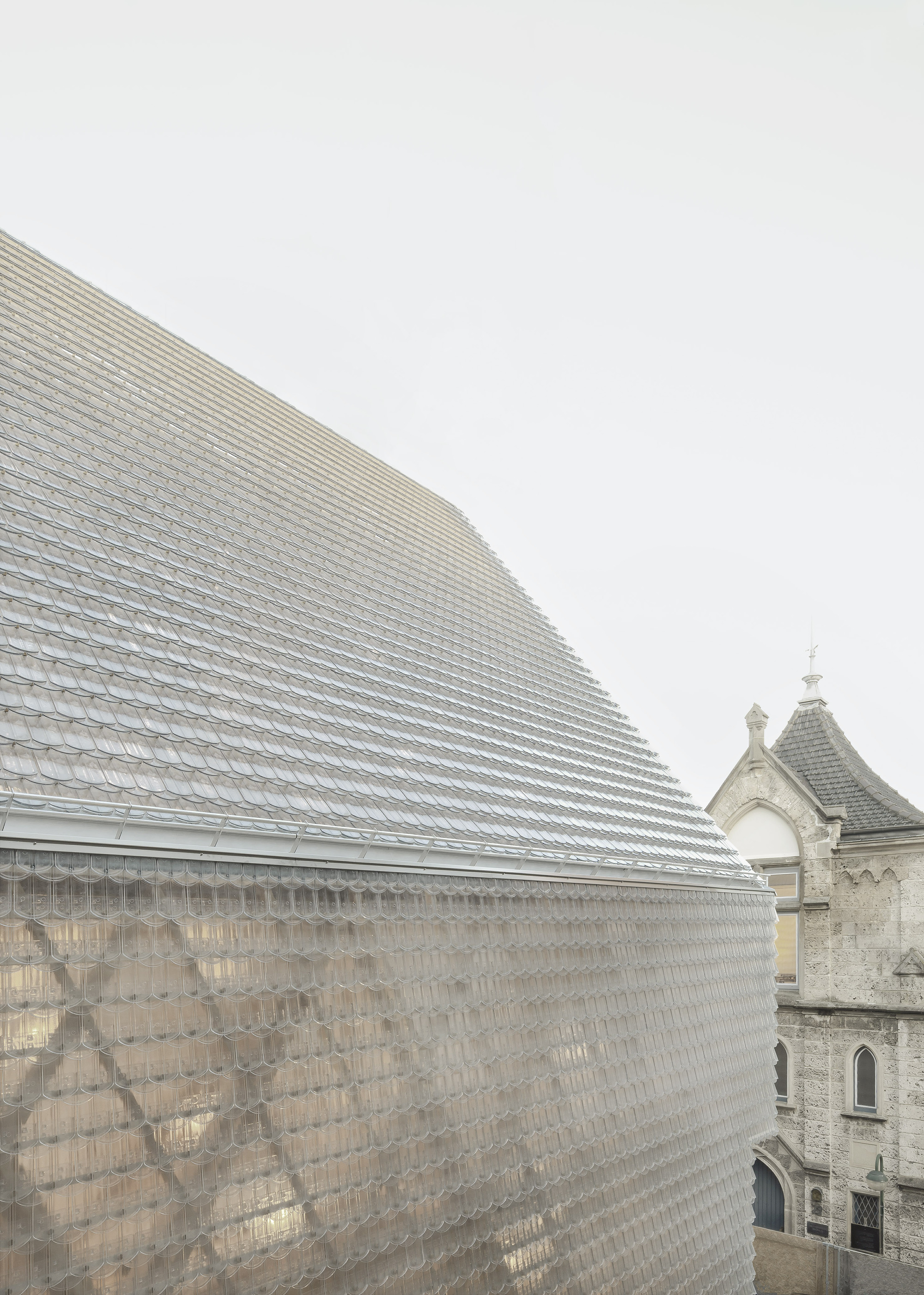Faceted glass tile roof volume adjacent to a historic stone church tower under grey skies