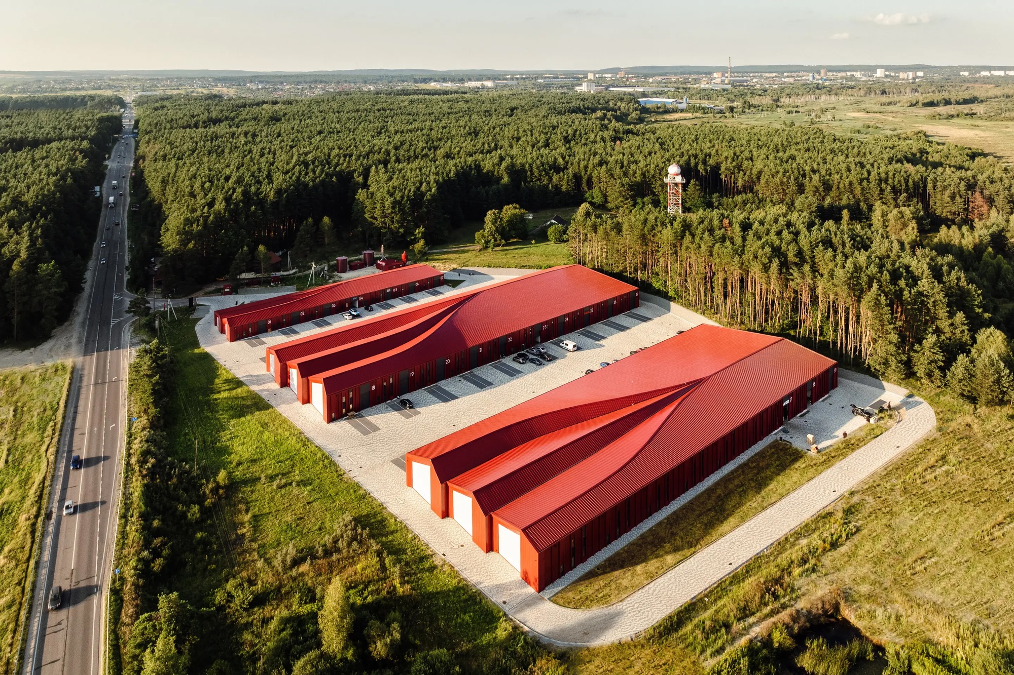 Aerial view of three red metal storage buildings with sawtooth roofs surrounded by forest and farmland