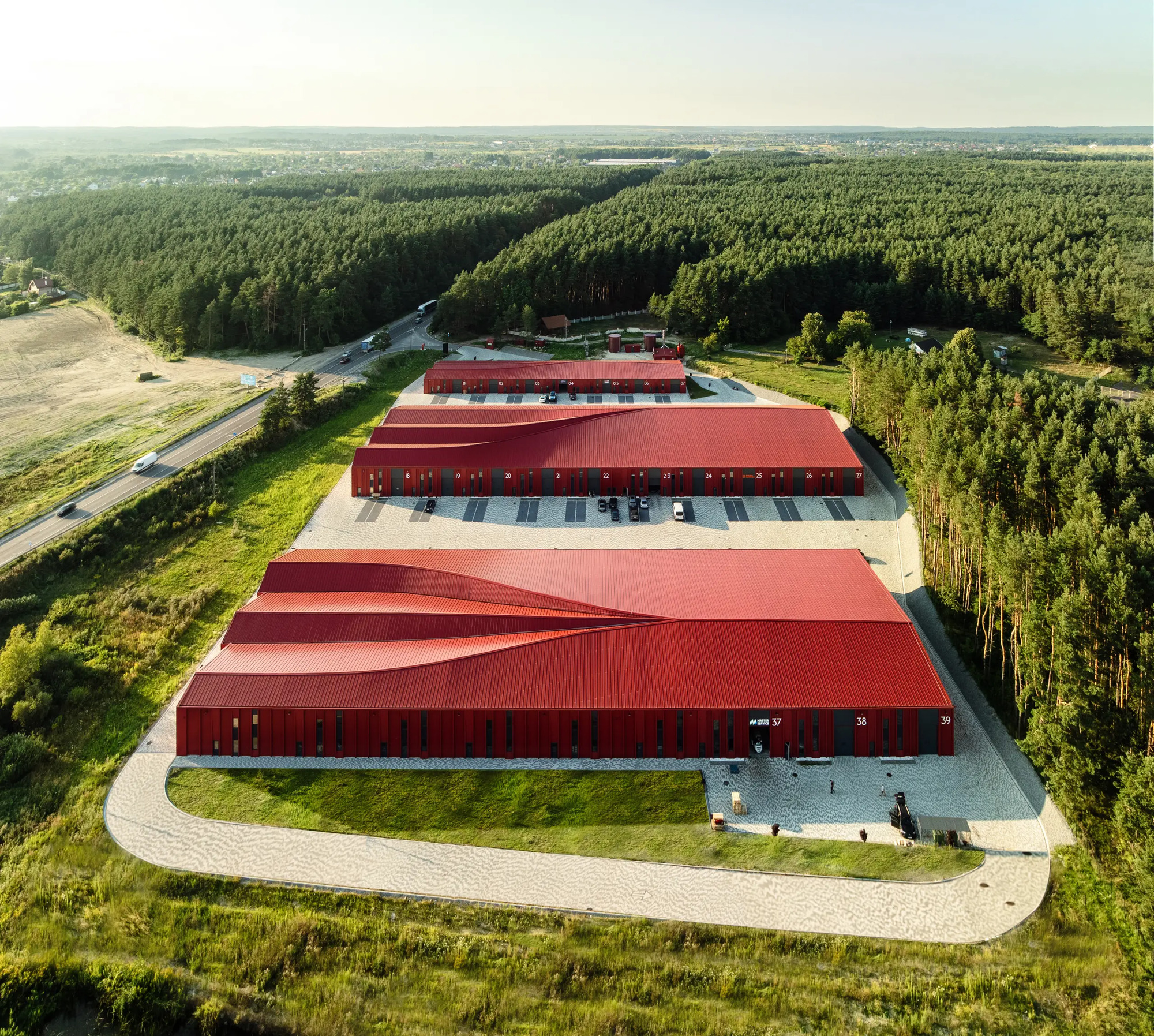 Drone view of three storage halls with folded red roofs on a curved site beside forest